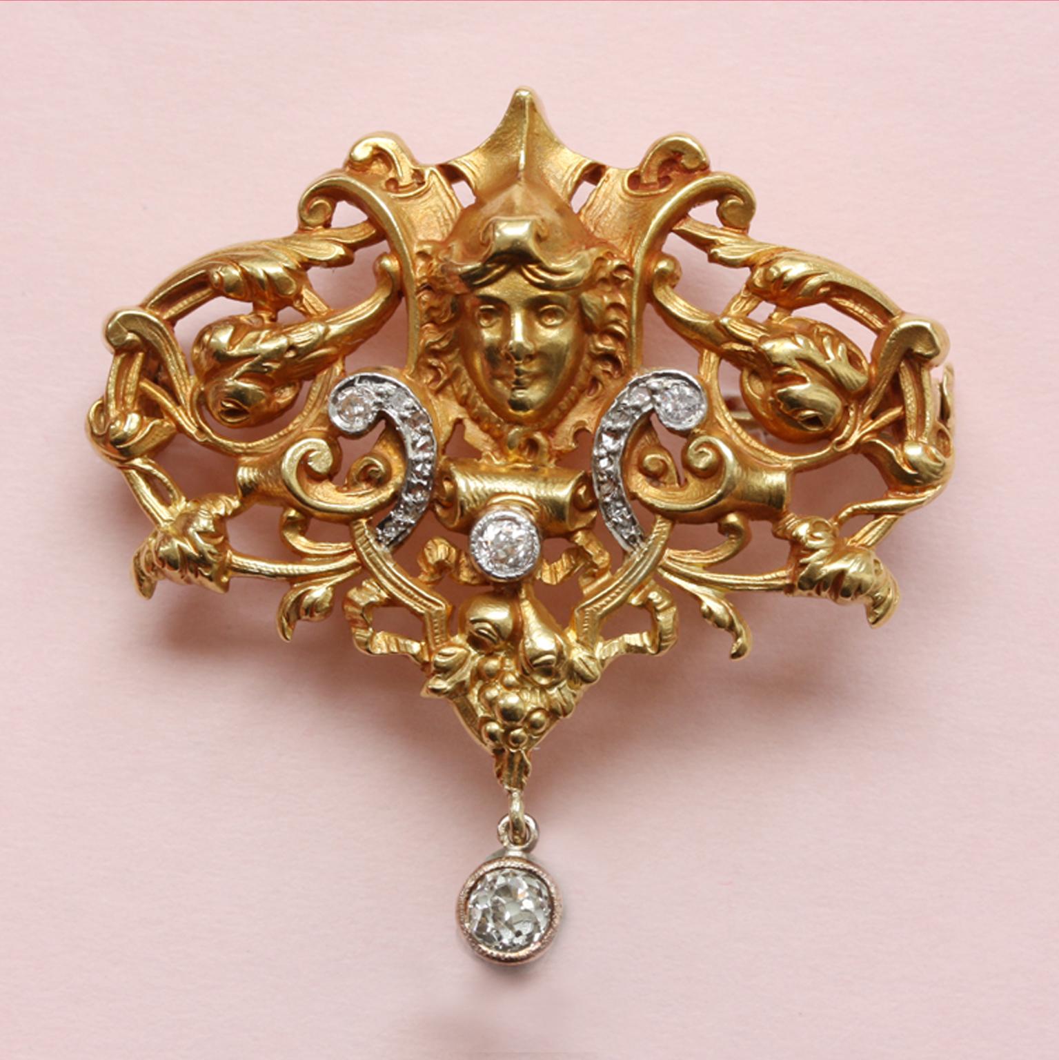 An 18-carat gold, openwork mascaron and scroll brooch centring a lady with an elaborate foliate and floral design, highlighted by two scrolls set with old-cut diamonds and a diamond drop, mille griffe set. Most likely France, circa 1890. The gold