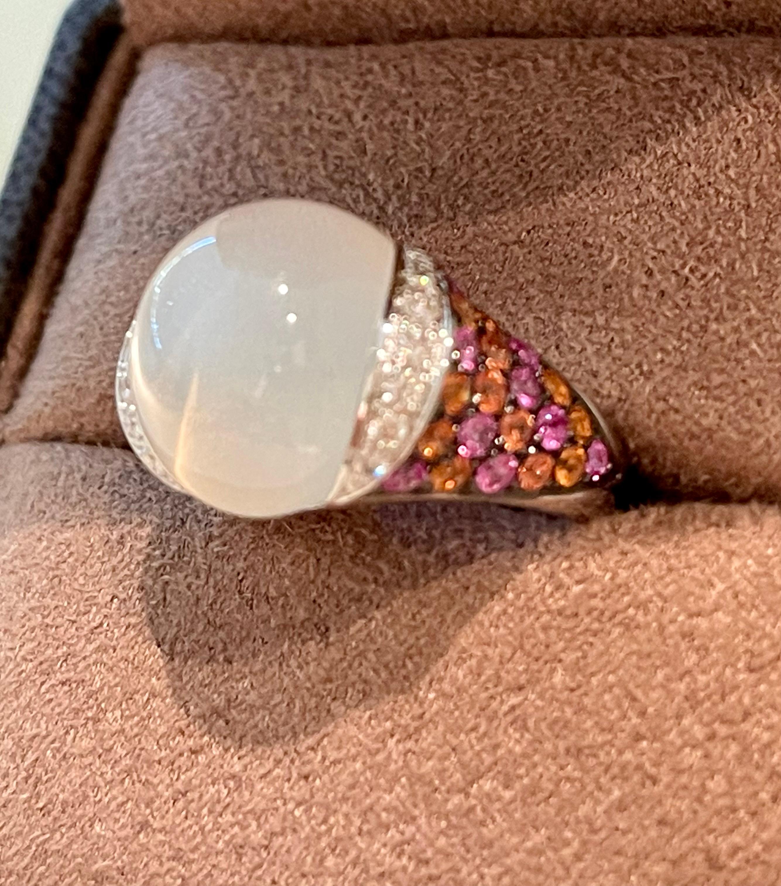 This gorgeous Ring crafted in 18 K white Gold features a white Moonstone Cabochon that dispays the charecteristic adularescence. Flanked by brillinat cut Diamonds, F color, vs clarity. The shank of the ring is pave set with orange and pink fancy
