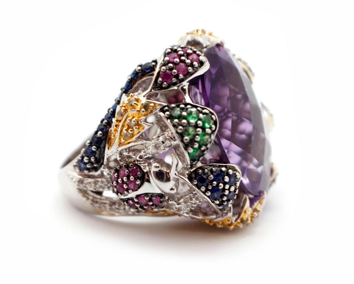This beautiful ring features a large, oval-cut amethyst set into 18k white gold. The amethyst weighs approximately 13.70cts. The shank of the ring is decorated with a variety of colored stones including green tsavorite, diamonds, and