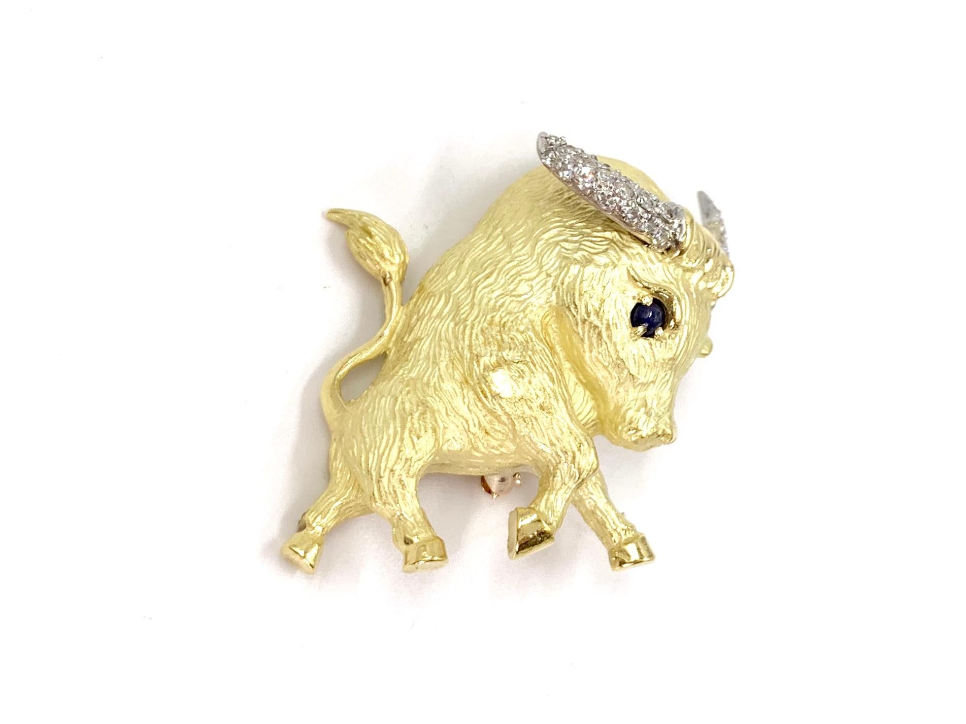 An adorable 18 karat yellow gold charging bull features a single dark blue cabochon sapphire and white single-cut diamond encrusted horns on this hand made brooch. Bull is hand finished with a matte/acid wash throughout the body and polished tail,