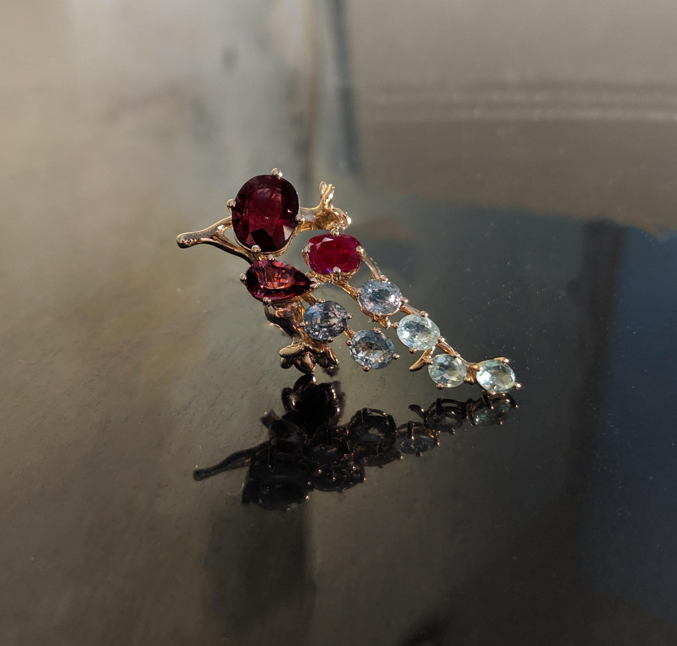 This Tobacco Flower contemporary brooch is made of  18 karat yellow gold.
The natural gems are:
Oval ruby 4,27 carats, dimension: 11,16x9,36 mm.
Oval cold red sapphire, 0,95 carats.
Pear cut cold malaya garnet, 1,25 carats.
Green oval cut sapphire,