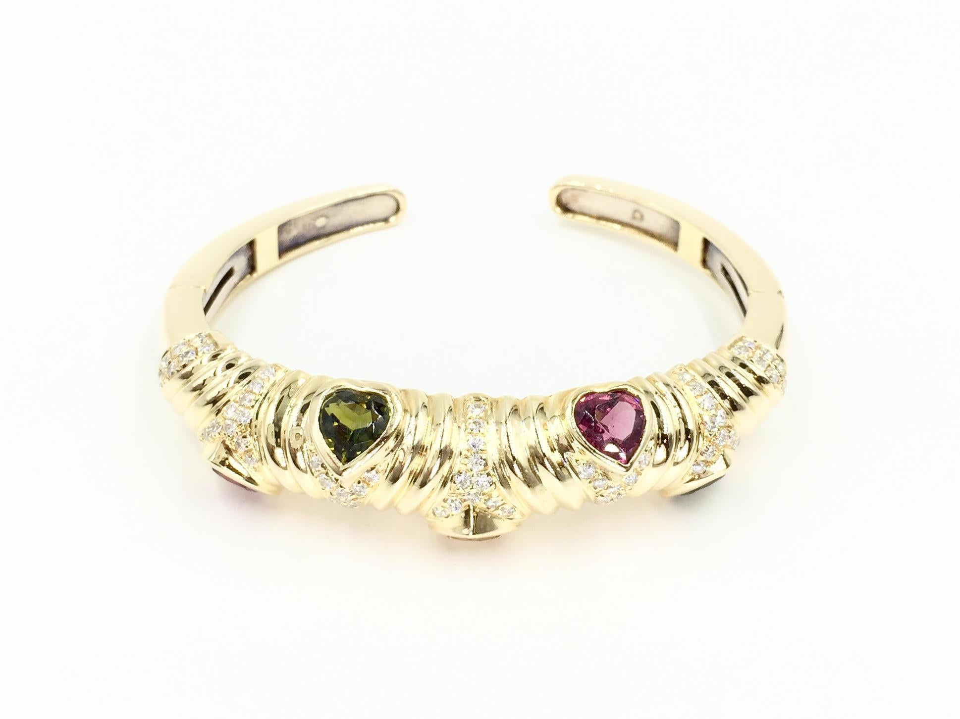 A solid and fashionable 18 karat yellow gold cuff bracelet featuring approximately 1.70 carats of round brilliant diamonds, approximately G color, VS2 clarity, and bezel set heart shape semi precious stones - two green tourmalines, two pink