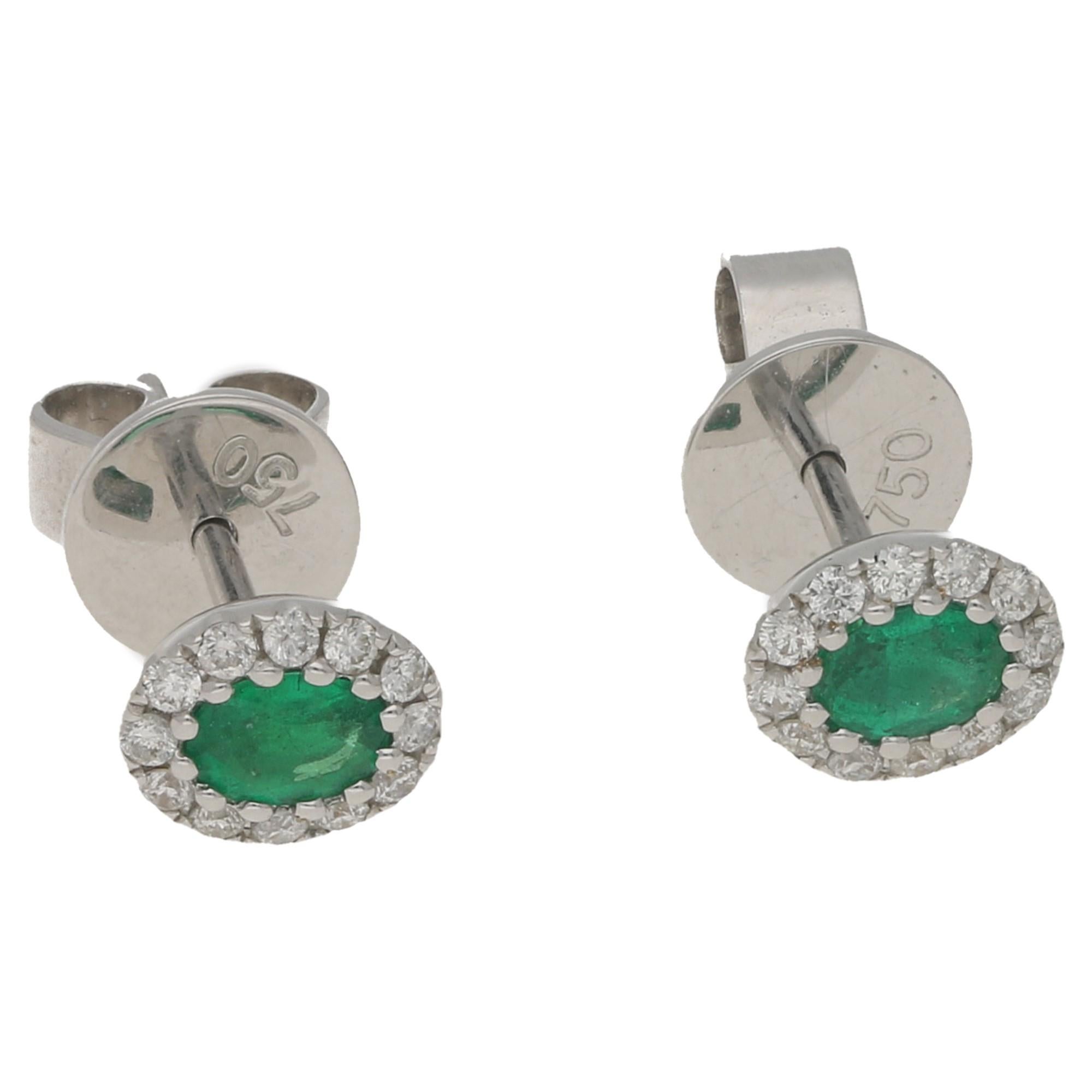 A pair of classic emerald and diamond oval cluster stud earrings. Set in 18ct white gold, the central emeralds totalling 0.27ct with excellent colour are surrounded by a cluster of round brilliant-cut diamonds totaling 0.15ct. Secured with a post