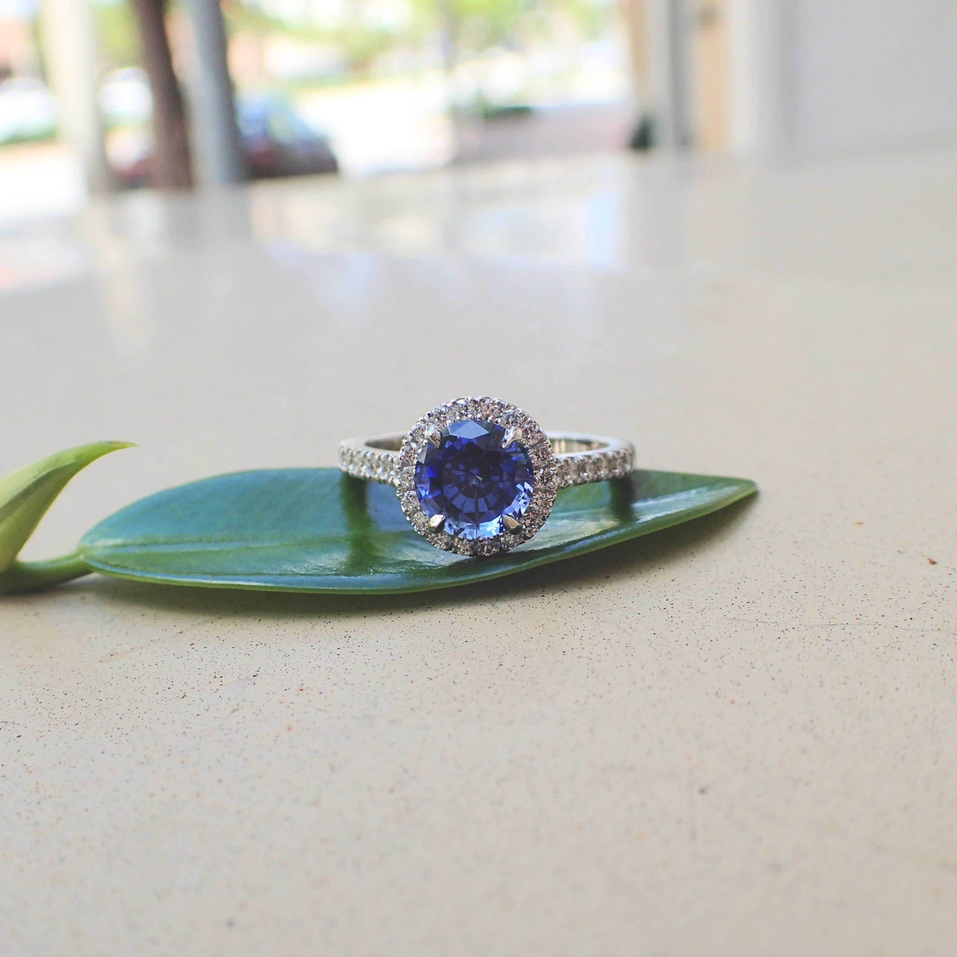 An 18k white gold ring is set with one (1) Round Brilliant Cut Chatham-Created Sapphire that measures 7mm x 7mm and weighs 1.85 carats with Clarity Grade VS-VVS and (18) Round Brilliant Cut Diamonds that measure 1.5mm x 1.5mm and weigh a total of