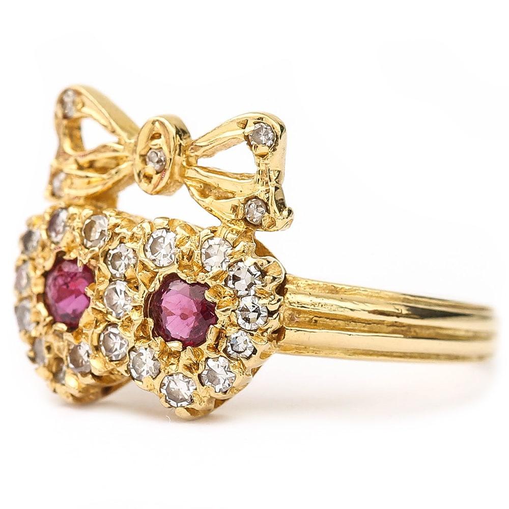 A pretty 18 karat yellow gold ruby and diamond sweetheart ring. The two entwinned hearts are set with two rubies, each 0.10cts, pave set with eight cut diamonds with an estimated diamond weight of 0.40cts. Set atop of the hearts is the tied diamond