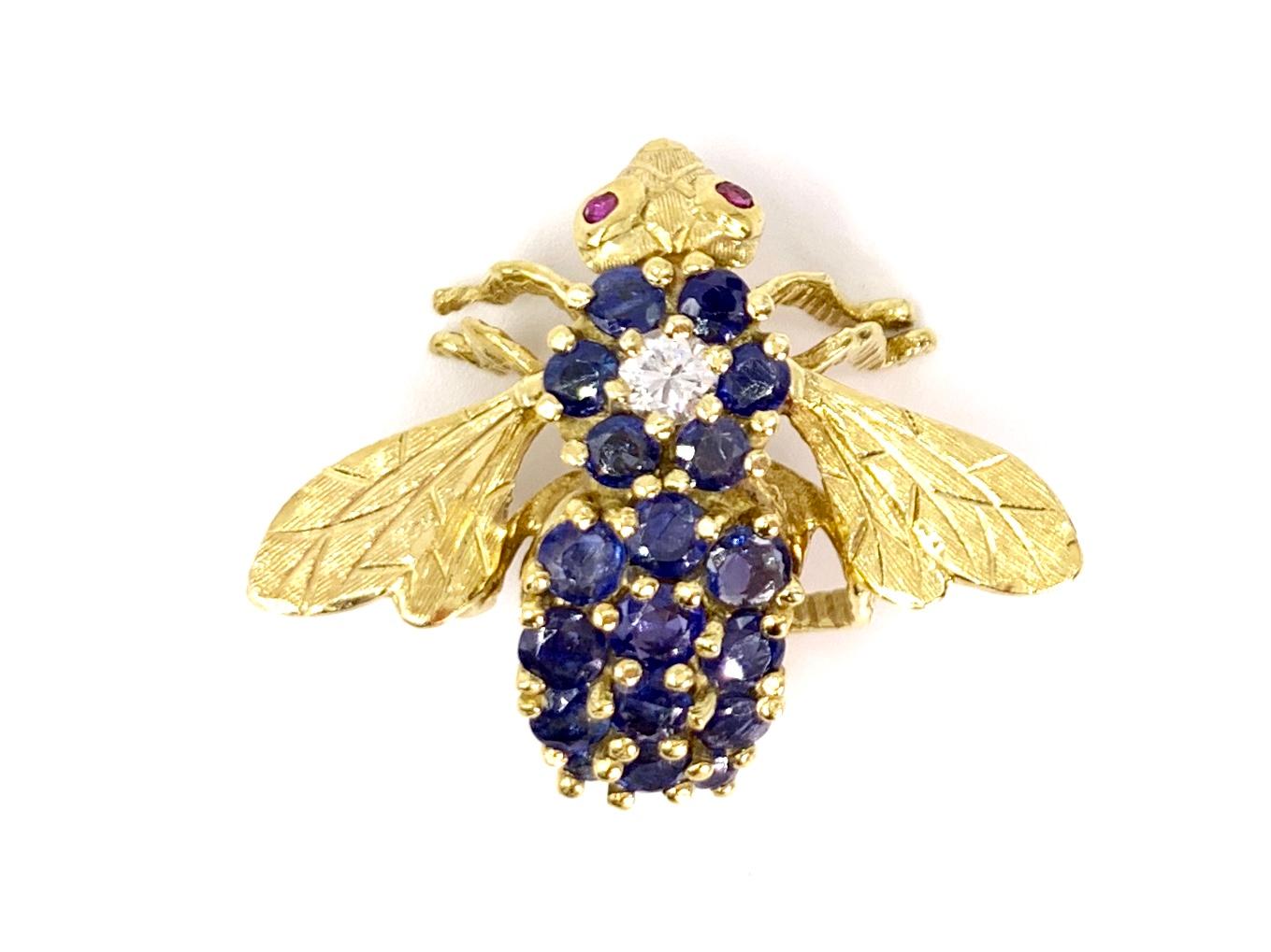 Adorable detailed flying bee brooch crafted by Hammerman Brothers in 18 karat yellow gold featuring one bright white diamond, 18 well saturated blue sapphires and 2 cabochon ruby eyes. Round brilliant diamond weighs approximately .20 carats at