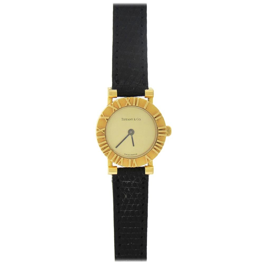 18K  yellow gold round women's Tiffany Atlas, the 19mm case with a  water resistant snap back, matte finish bezel with polished embossed gold roman numeral hour markers and blacked steel baton hands, champagne dial, quartz movement. 