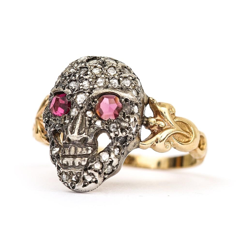 An 18 karat yellow gold, silver set ruby and diamond skull ring in the memento mori style. The ruby eyes and diamonds are rose cut (an early form of diamond/stone cutting where the base of the stone is flat and the top is pointed). The skull