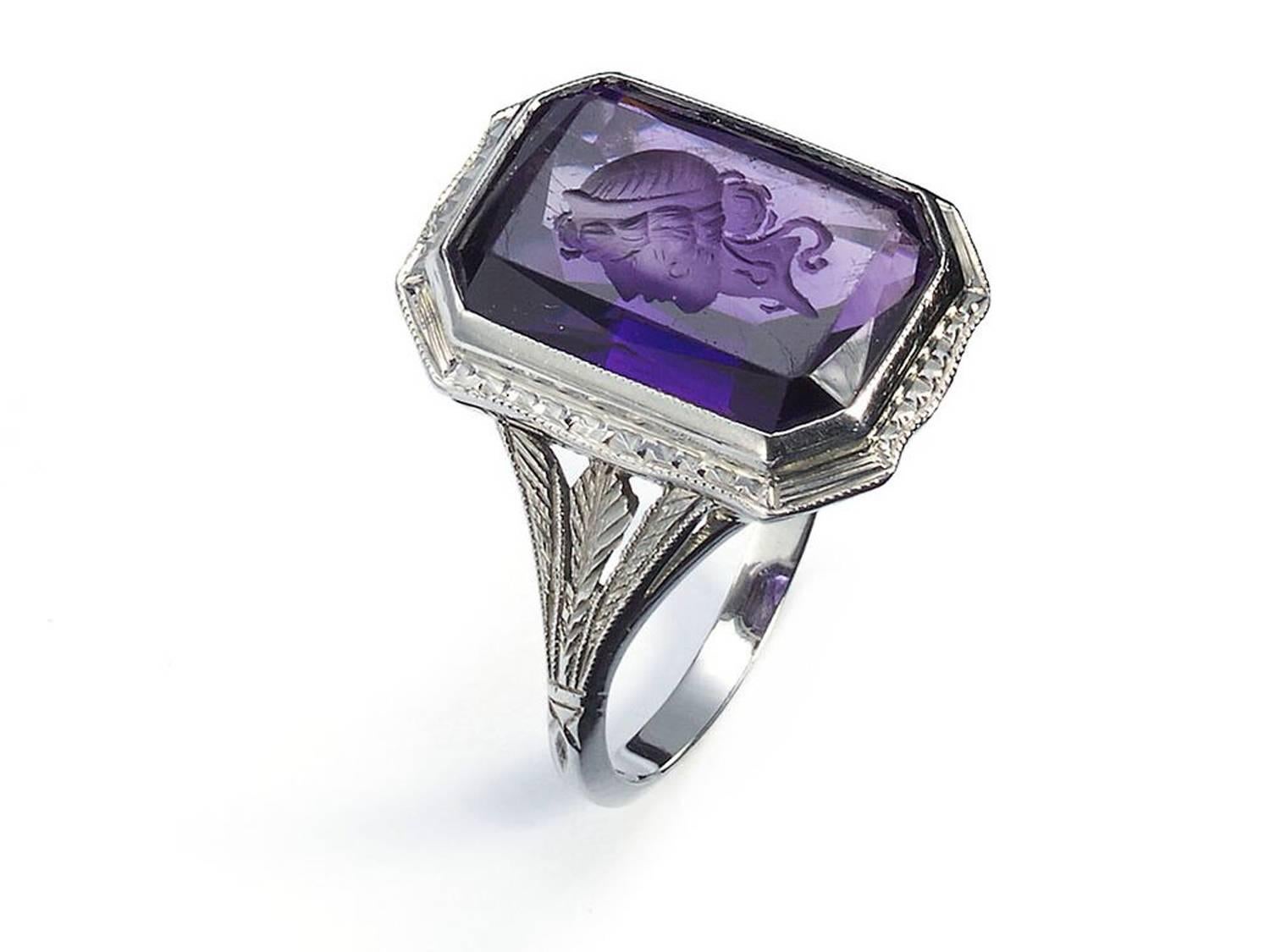 An amethyst intaglio ring, with an emerald-cut amethyst, carved with the head of a Roman style woman, in a millegrain edged rub over setting, with a square nailhead design and millegrain edged surround, with carved and millegrain trefoil shoulders,