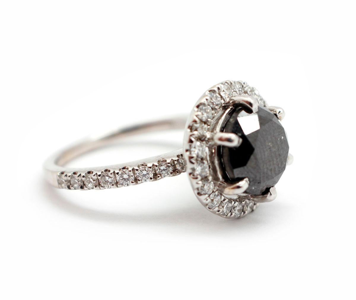This ring is made in 18k white gold, and it holds a 3.45-carat, round brilliant-cut black diamond at its center. The black diamond is accompanied by a GIA/JGL report, #20181. The ring features a white diamond halo and white diamond accents down the