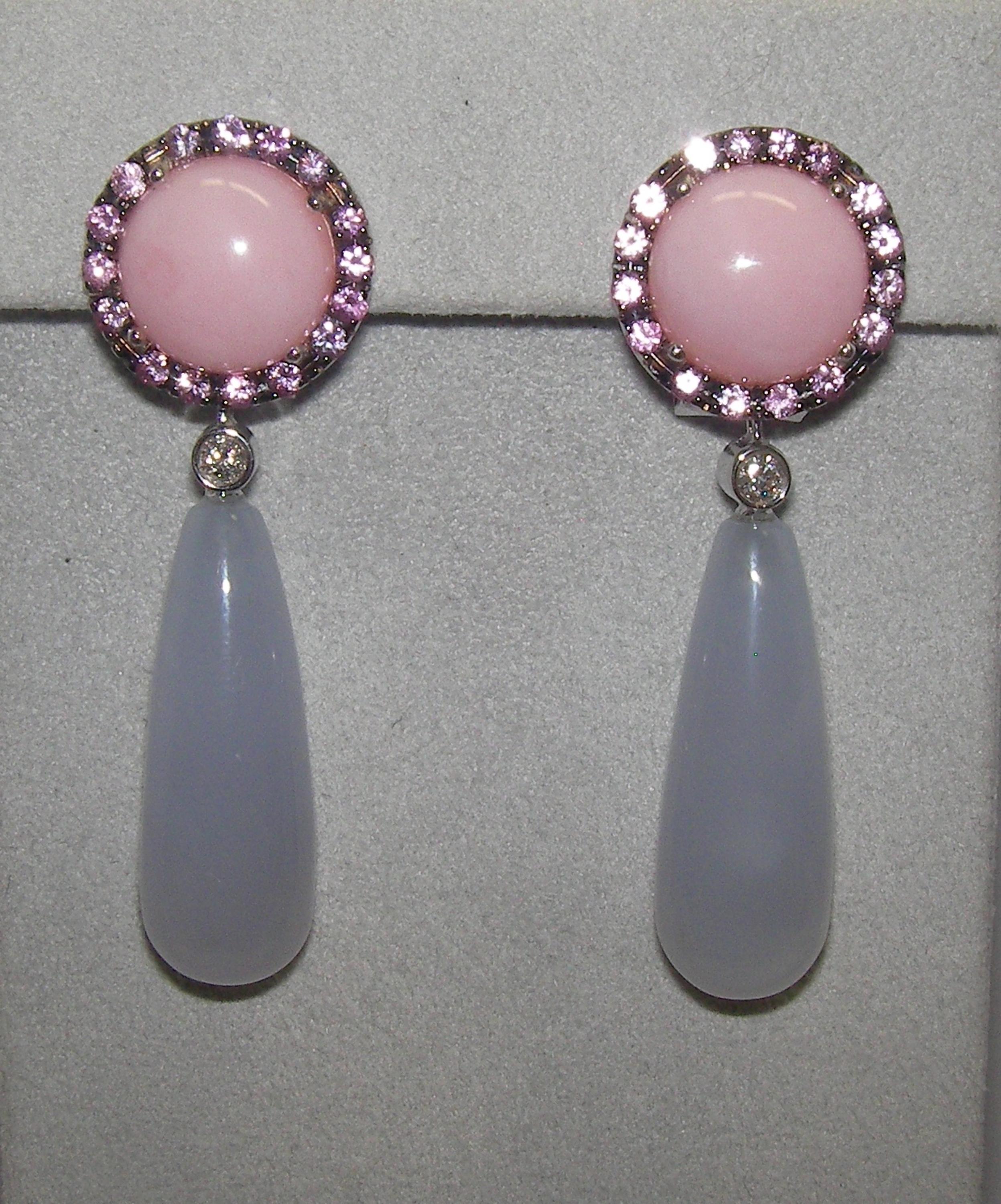 These 18 Karat White Gold Chalcedony Diamond Opal and Sapphire Drop Earrings feature a halo of rose colored Sapphires and Diamonds around a round cabochon Rose Opal center stone. The base is then followed by a hanging pear shaped cabochon, separated