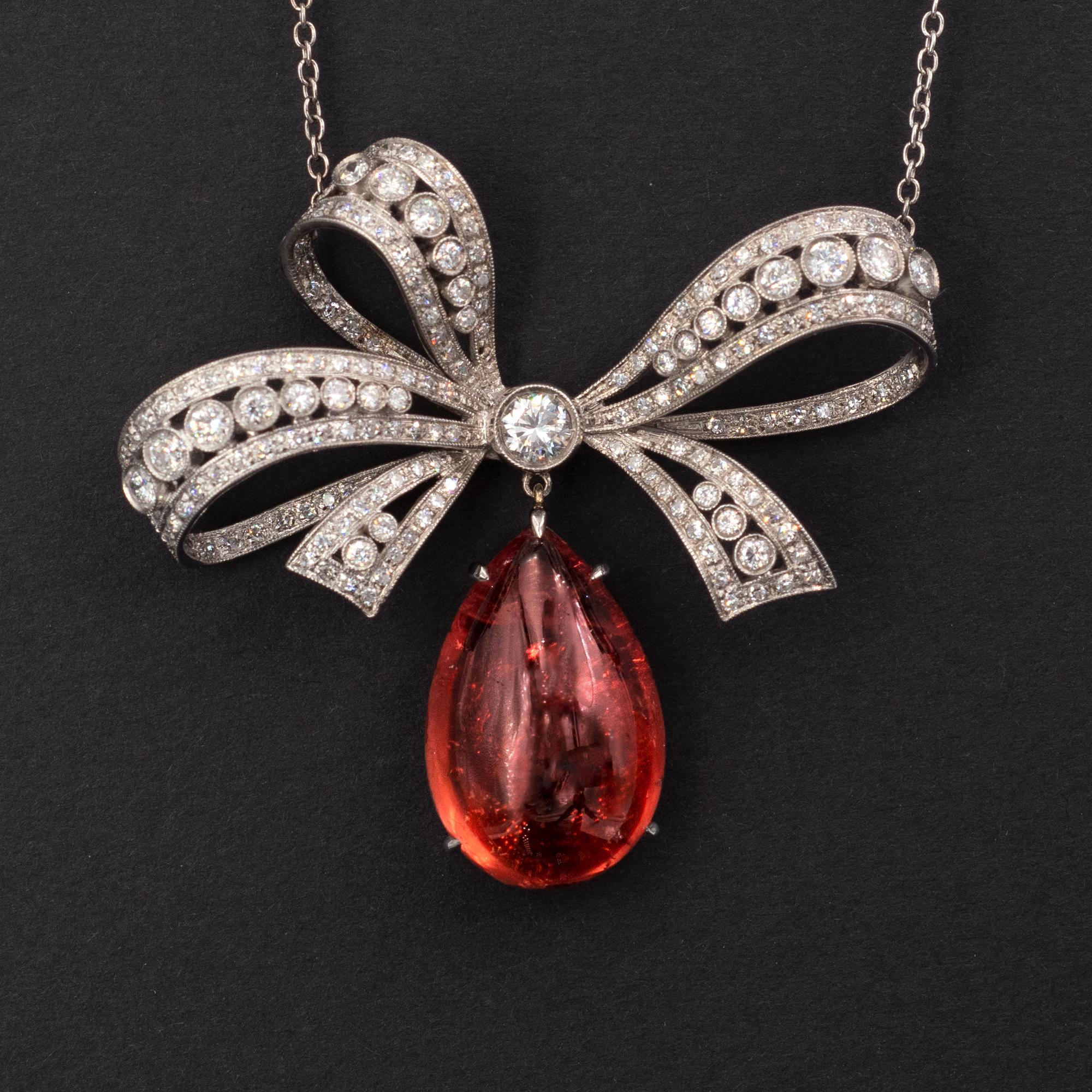 This is a refined necklace: a feminine bow with a sophisticated work reminiscing the Edwardian Belle-Epoque era, set with white diamonds, from which dandle a striking, pear-shaped cabochon tourmaline displaying a radiant pinkish-orange color. The