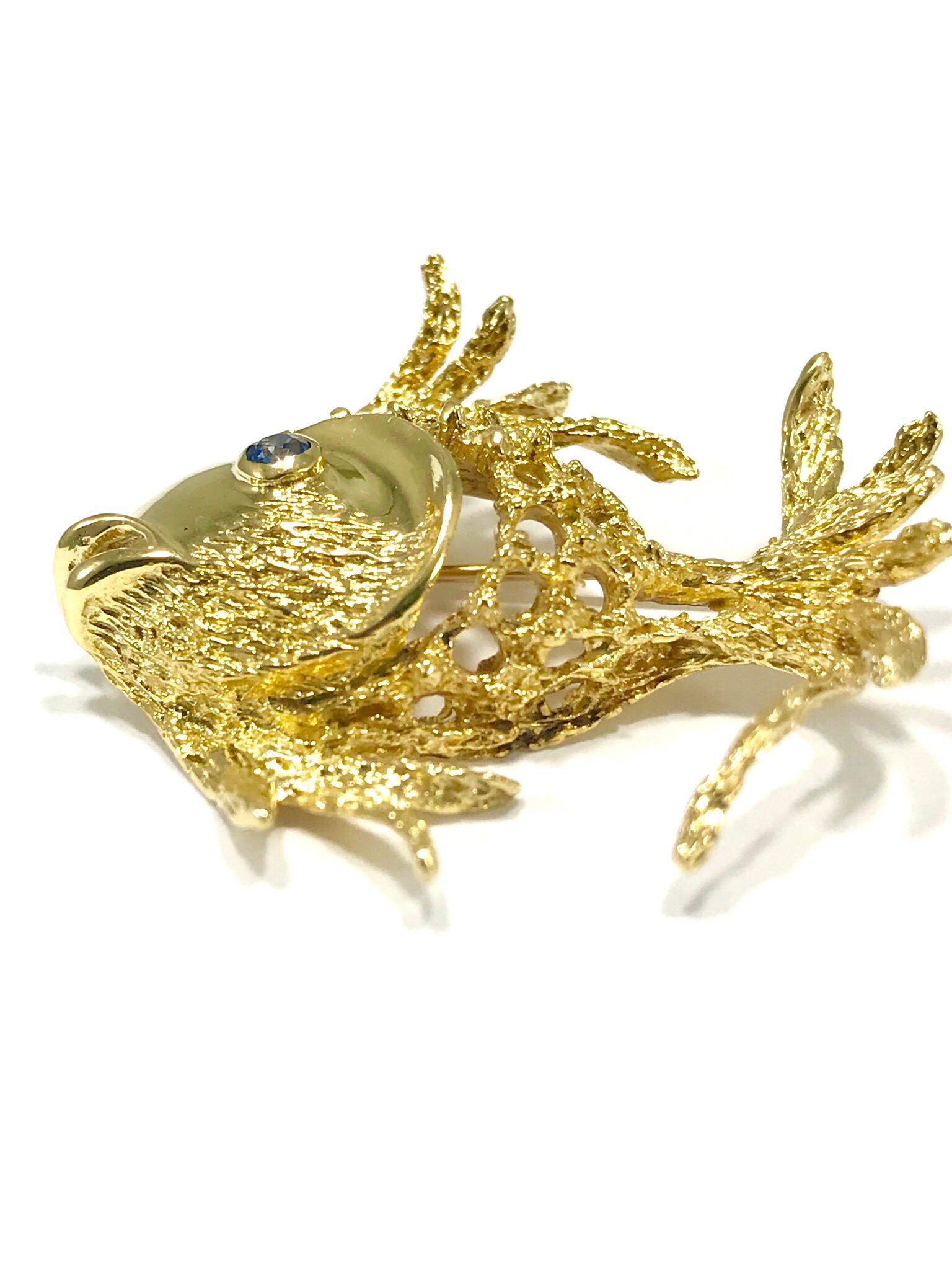This is a whimsical 18 karat yellow gold angel fish brooch, with a single sapphire eye.  The sapphire is 0.15 carats, bezel set.  The fish body is made up of textured open work gold.  The pin features a a single pin closure.  The tail fin is stamped