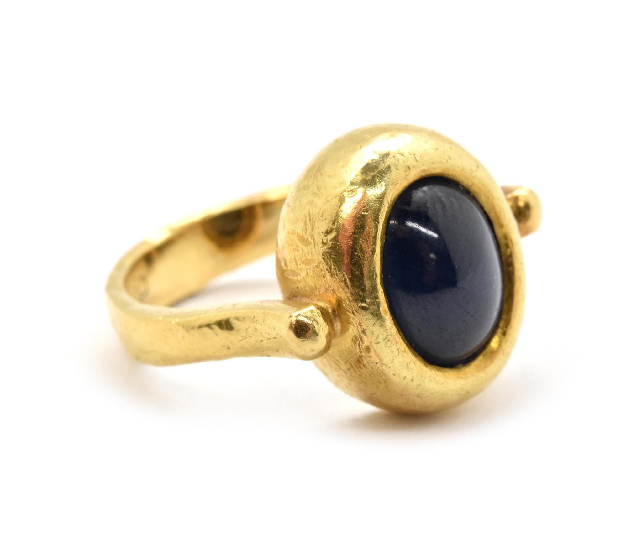 This ring is made in solid 18k yellow gold and it features a cabochon blue sapphire at its center. The semi-opaque deep blue sapphire is bezel set in a chunky bezel with a lightly hammered texture. The shank of the ring is narrow and attaches at the