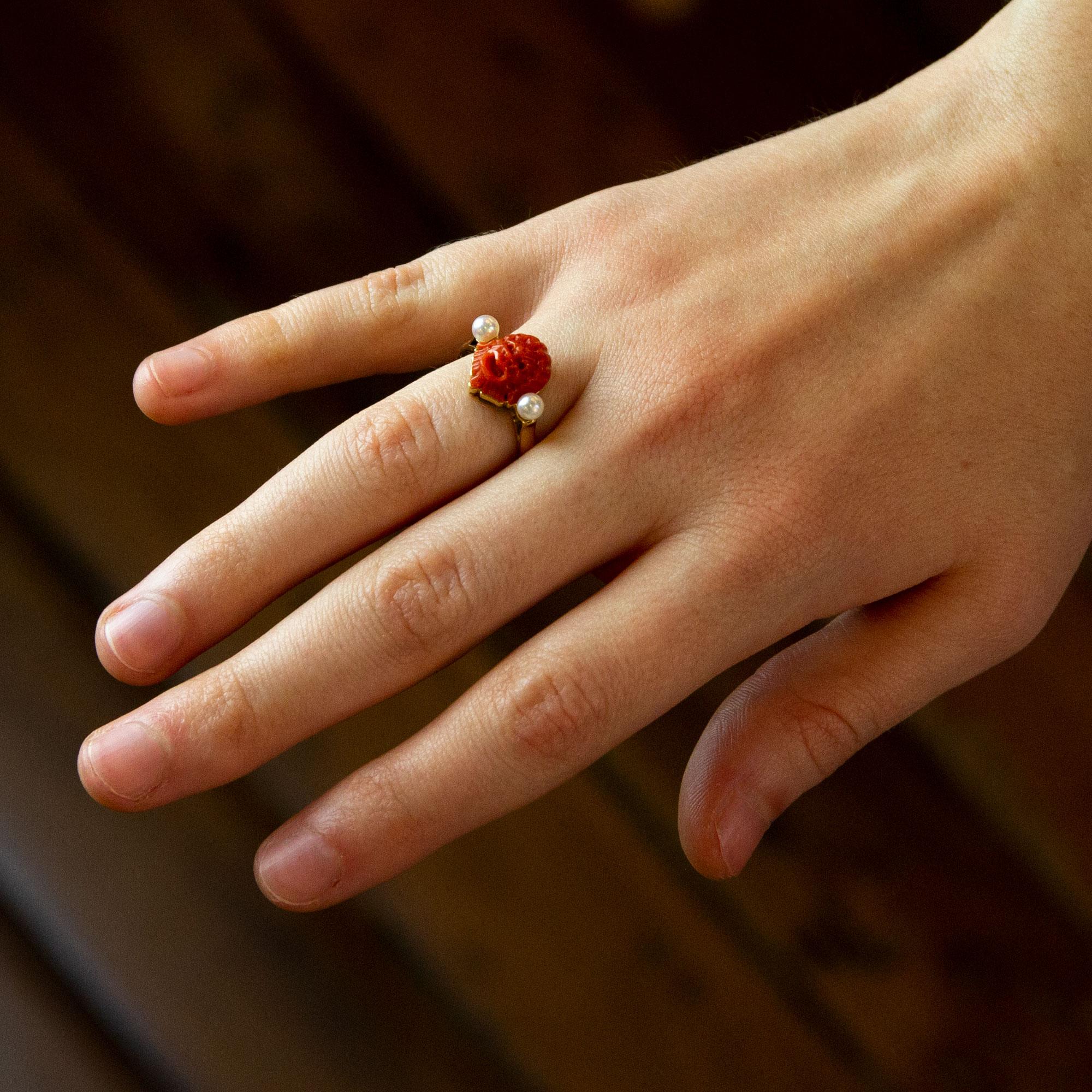 A fun carved dark orange coral and cultured pearl ring crafted from 18 karat yellow gold. The central piece of coral depicts a carved monkey head which is flanked by a pair of cultured white pearls measuring 3.75mm. The shank of this ring is very