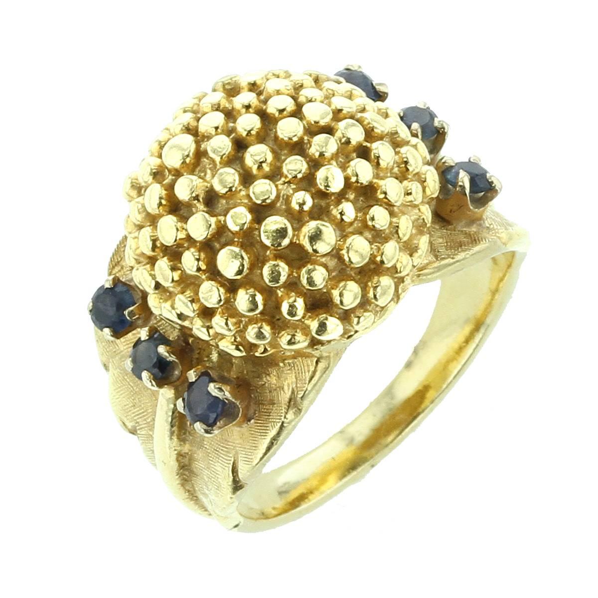 A unique ring composed of a centered cluster of granulation flanked by six round blue sapphires, measuring approx. 2.50 mm. It consists of a massive, high dome form with sculptured and texture shoulders with a half-round shank, 18K Yellow Gold. Size