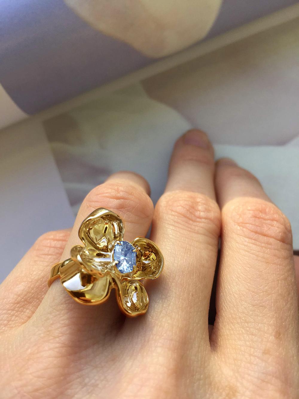 This Magnolia Flower contemporary engagement or cocktail ring is made of 18 karat yellow gold and features a clear and shiny light blue sapphire weighing 0.65 carats. The water-like surface of the gem multiplies the light, mirroring on the golden