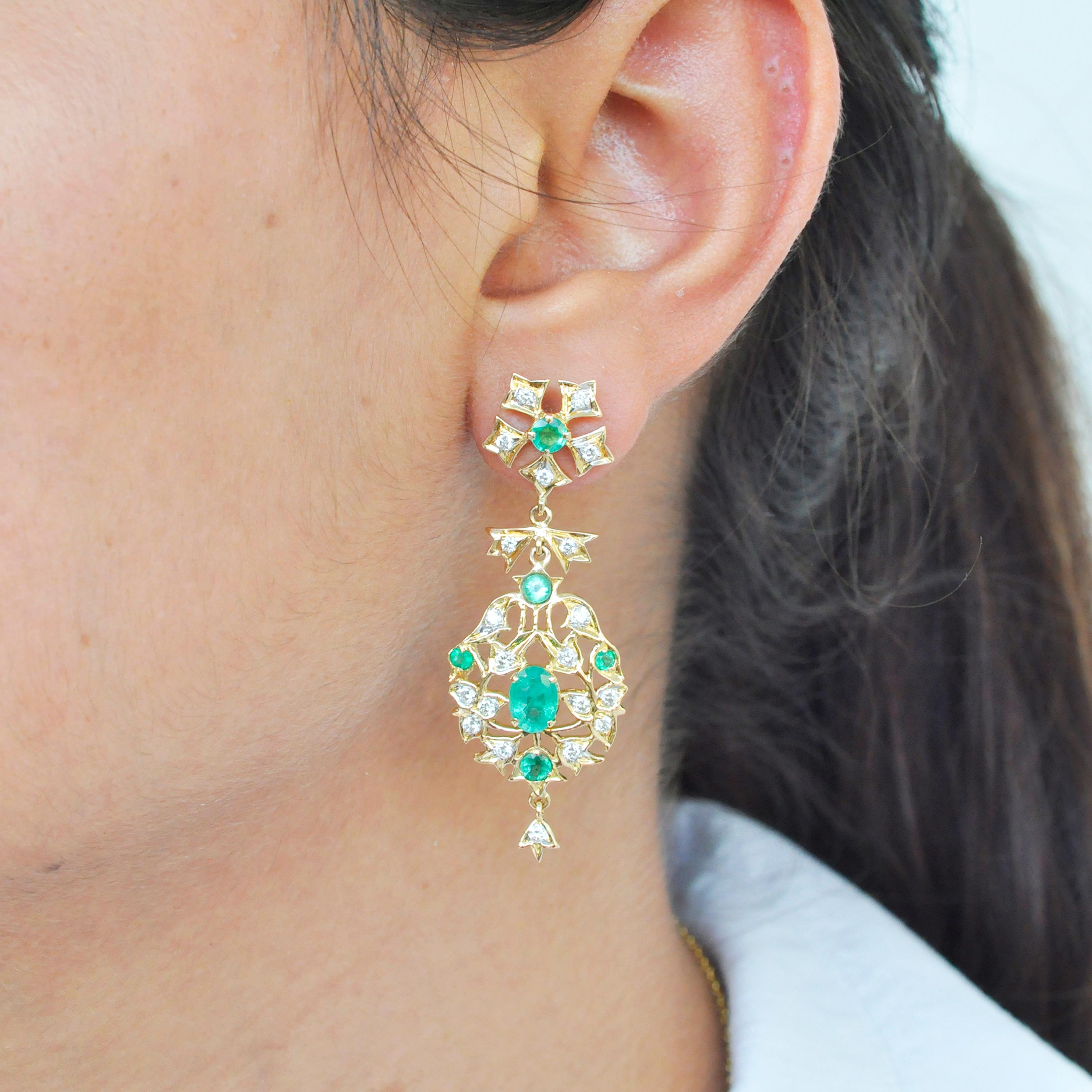 These beautiful emerald diamond dangle earrings are made in 18K gold. A mix of high quality round and oval emerald gemstones are placed in between a delicate ensemble of diamonds set in tulip motifs of gold. If you are looking for something chic and
