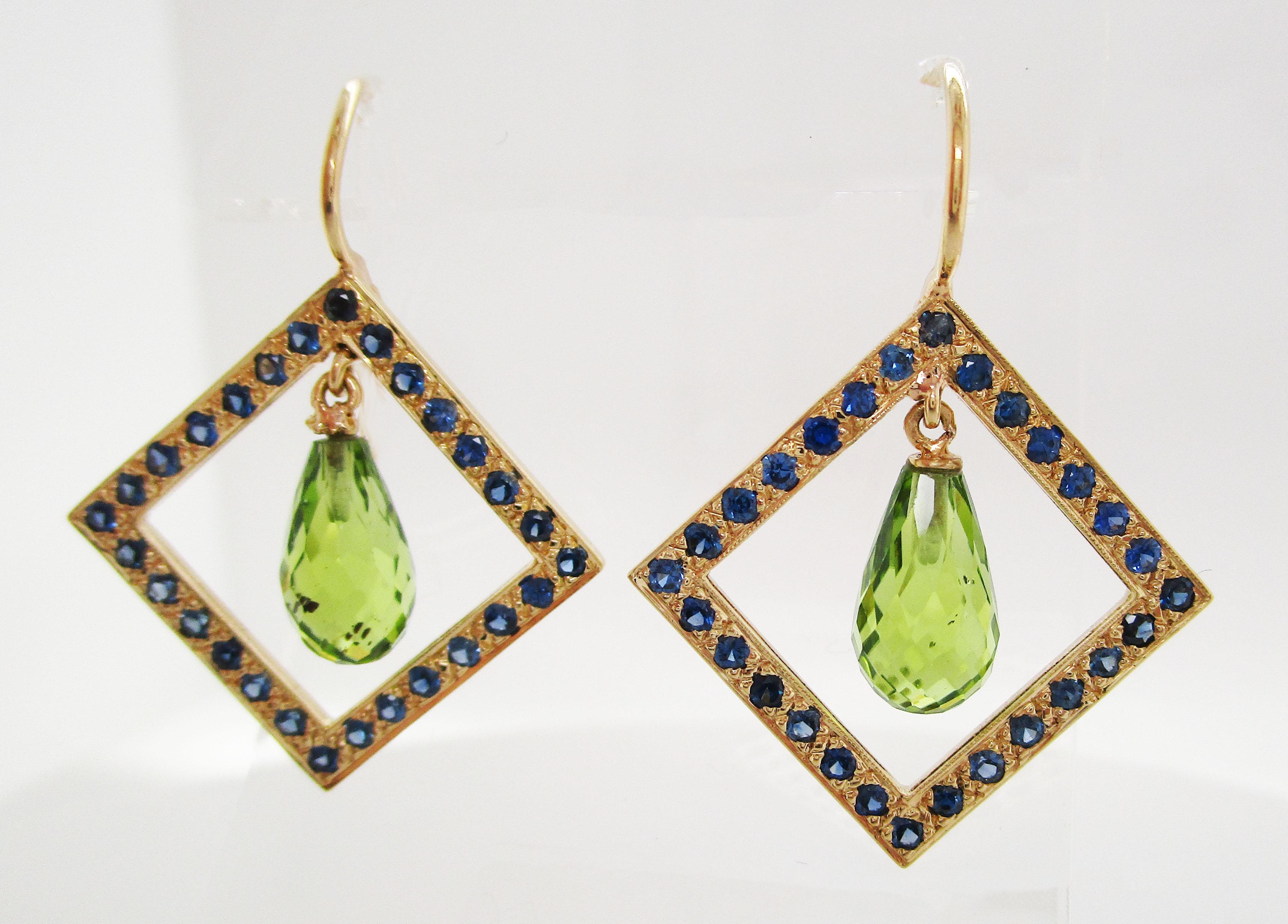 This is a fantastic pair of drop earrings in 18k yellow gold with a unique geometric design featuring a frame of bright blue sapphires and a gorgeous peridot drop center. The square design frames the suspended briolette peridot center to create a