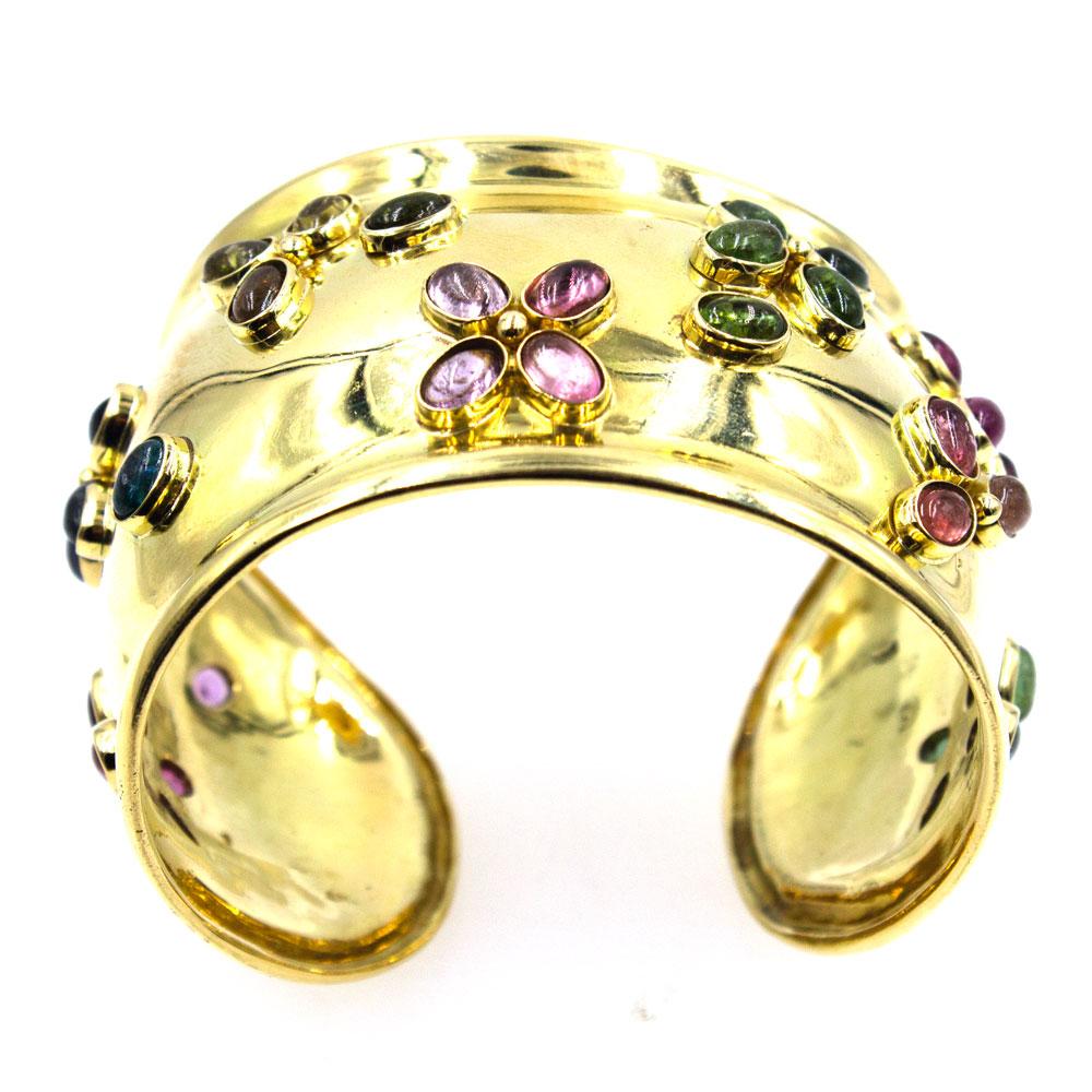 Wide colorful cuff bracelet in 18 karat yellow gold. The cuff features bezel set cabochon gemstones including rubelite, peridot, and blue topaz. The bracelet measures 1.75 inches in width and will fit up to a 7.0 inch wrist. 