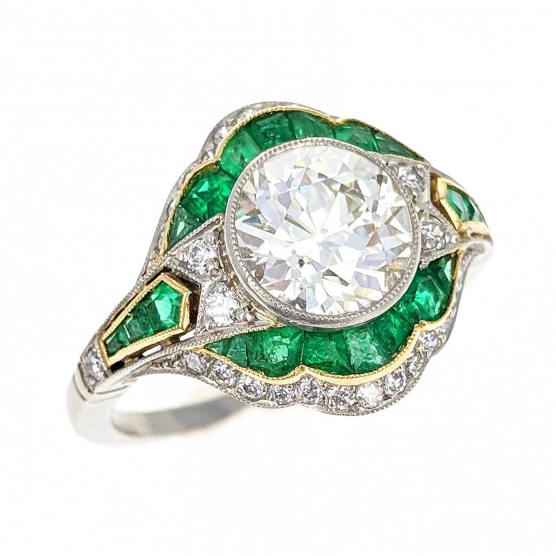 This gorgeous Art Deco style engagement ring centers upon a bezel set old European-cut diamond of 1.80 carats and estimated I/J color and VS clarity. It is encircled and flanked by pave set single-cut diamonds and calibre-cut emeralds set in yellow