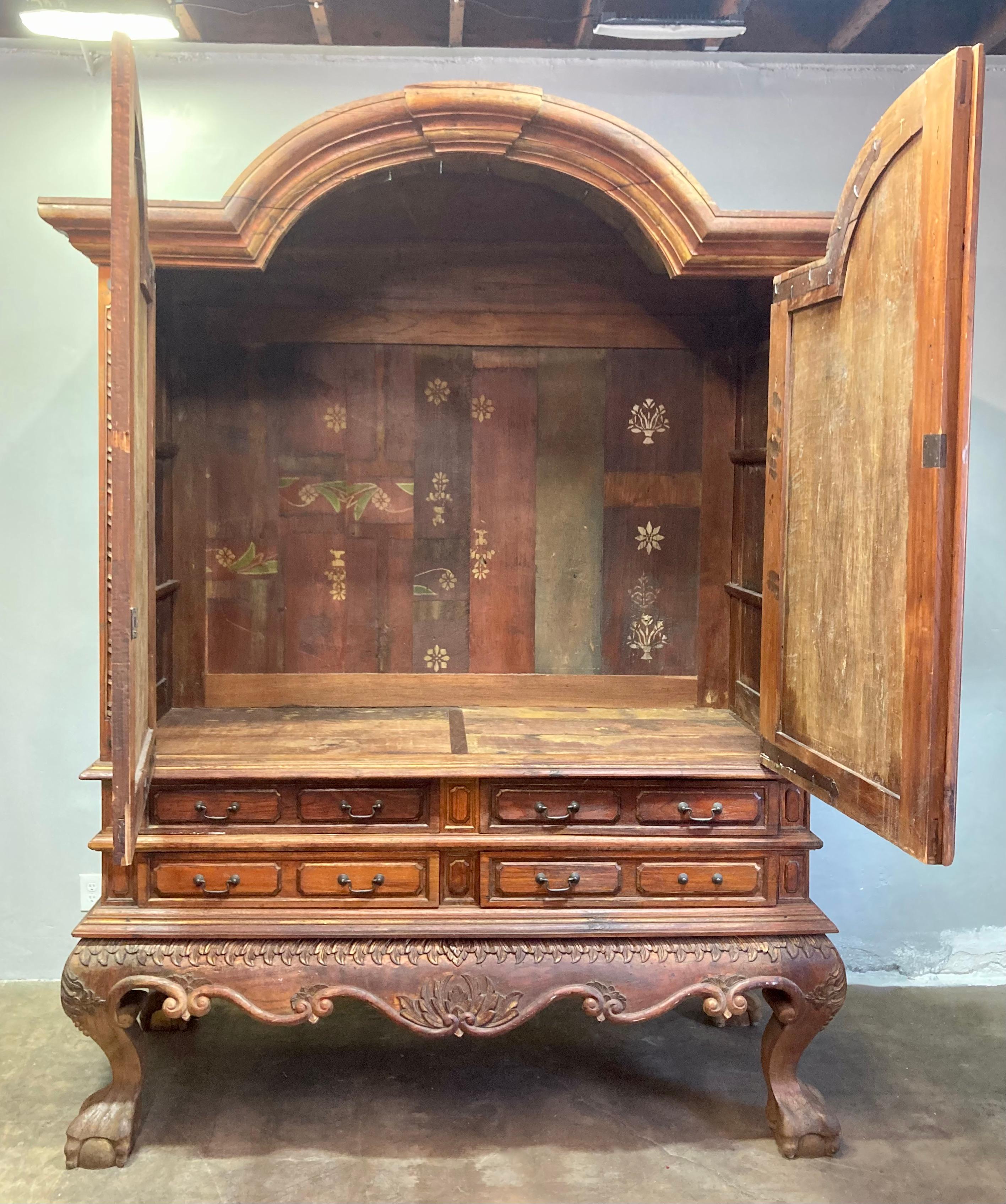 Precioso armario antiguo de estilo colonial holandés procedente de Indonesia. Sus dos estantes y 4 cajones ofrecen un almacenamiento espacioso. Los adornos de follaje de madera bellamente trabajados recorren los lados, los herrajes y detalles de