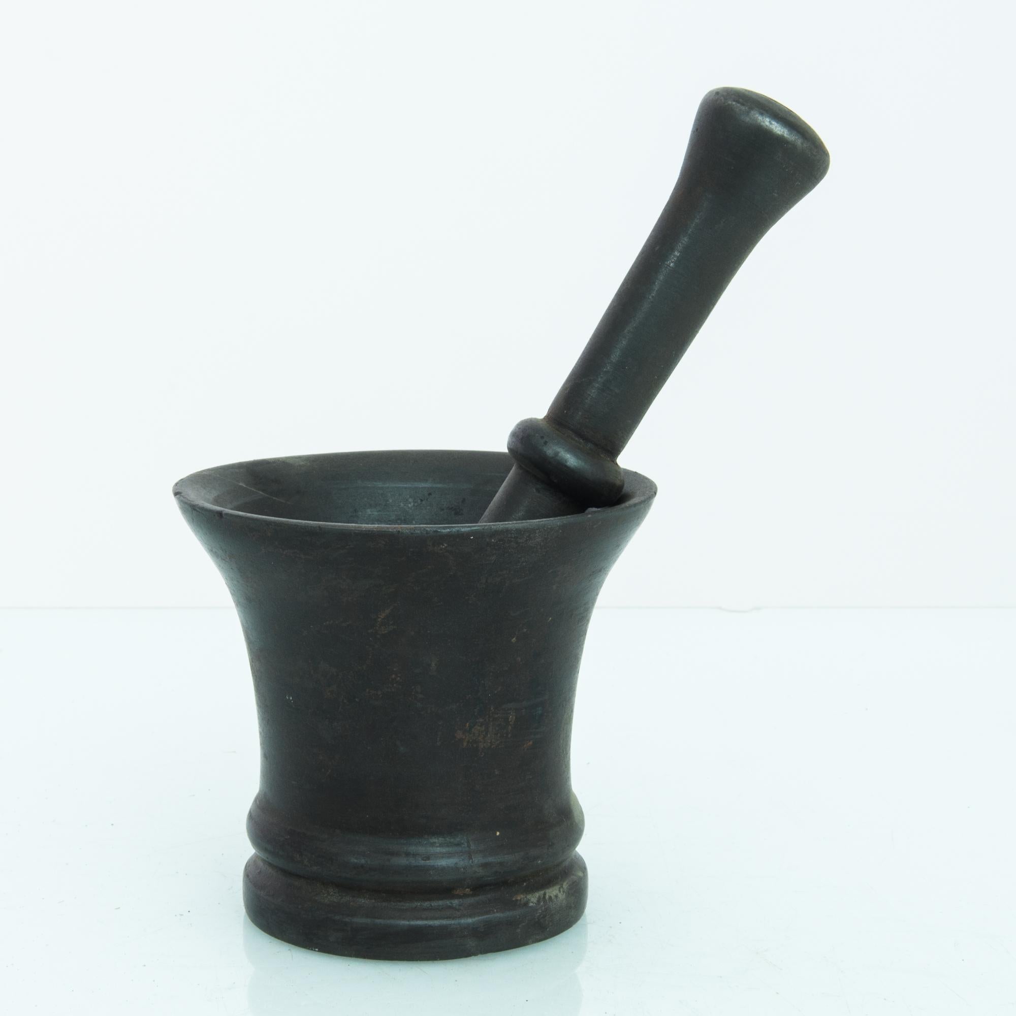 A cast iron mortar and pestle from Belgium, circa 1800. An upturned bell-shape in dark metal, accompanied by a long pestle with softly rounded ends. The surface of the metal has an enigmatic, multi-tonal quality, inflected with shades of indigo and