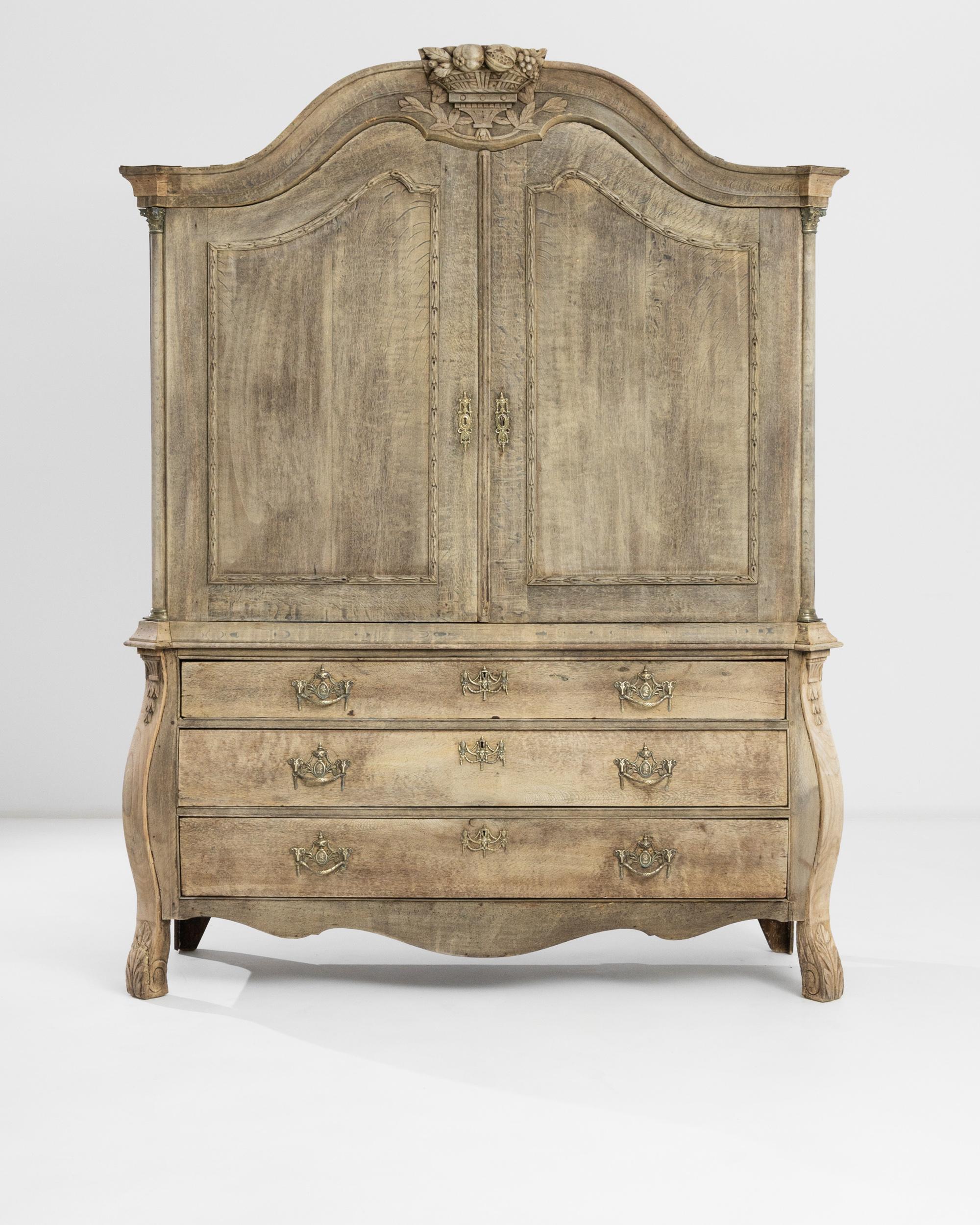 An 1800s Dutch bleached oak cabinet. This grand cabinet offers three sliding bottom drawers and a large top compartment fit with subsequent, smaller drawer pulls. A scrupulously hand-carved detail of a cornucopia sits atop it, while roman motifs