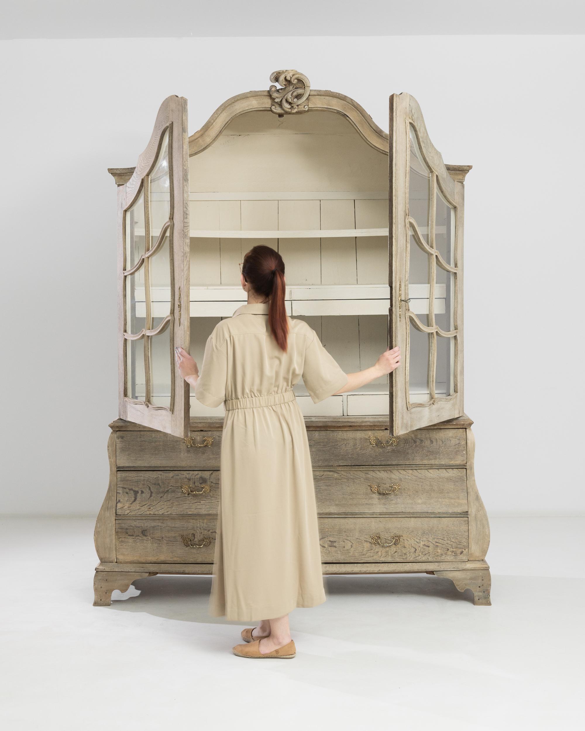 The silhouette of this antique oak vitrine evokes the lavish beauty of the Baroque. Built in the Netherlands circa 1800, serpentine drawers are crowned by an arched cabinet. The panes of the mullioned glass descend in delicate lace-like cascades to