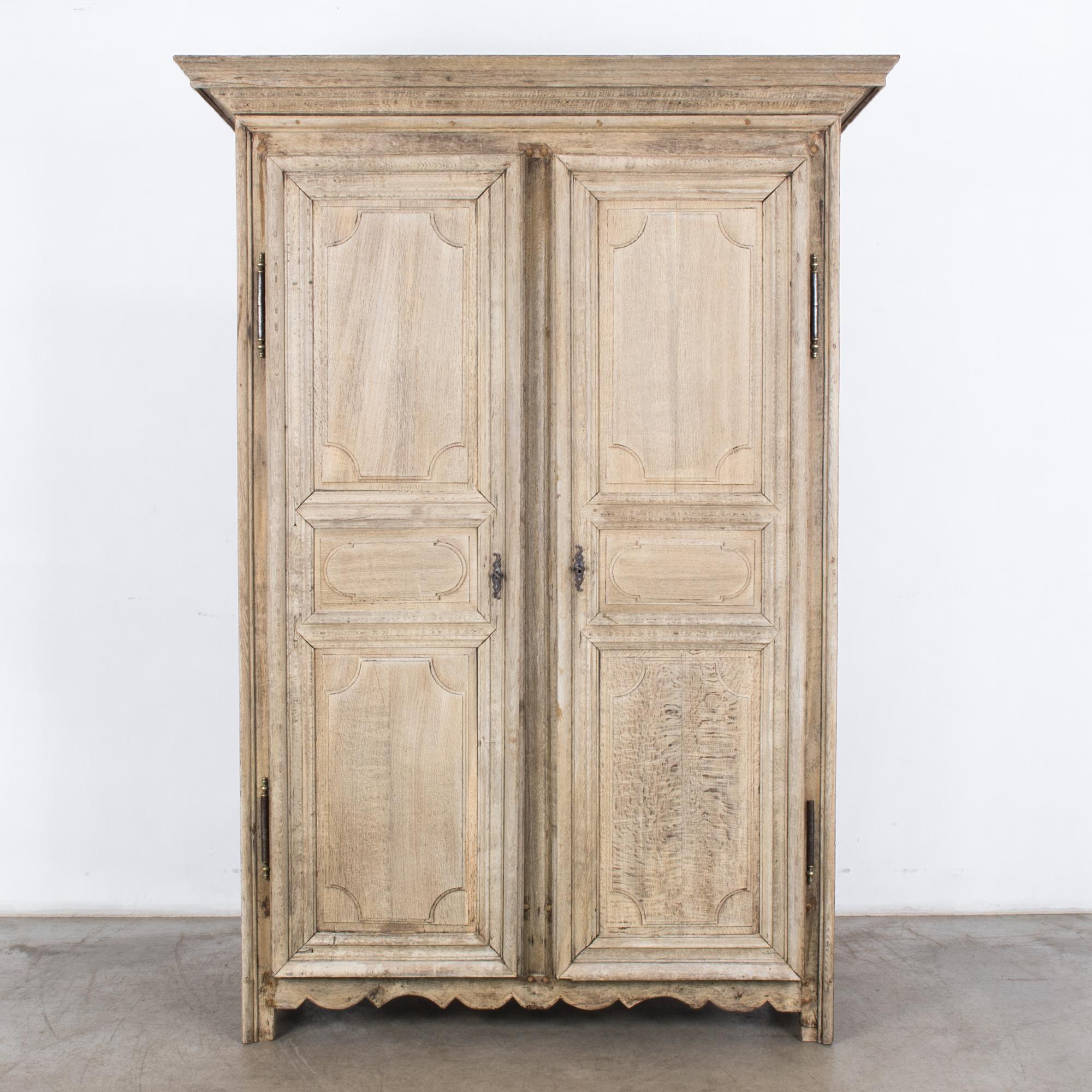 A bleached oak armoire from France, circa 1800. Paneled doors open into two interior shelves and a pair of lower drawers with golden handles. The wood has been restored to a bleached oak finish, highlighting the texture of the grain. The scalloped