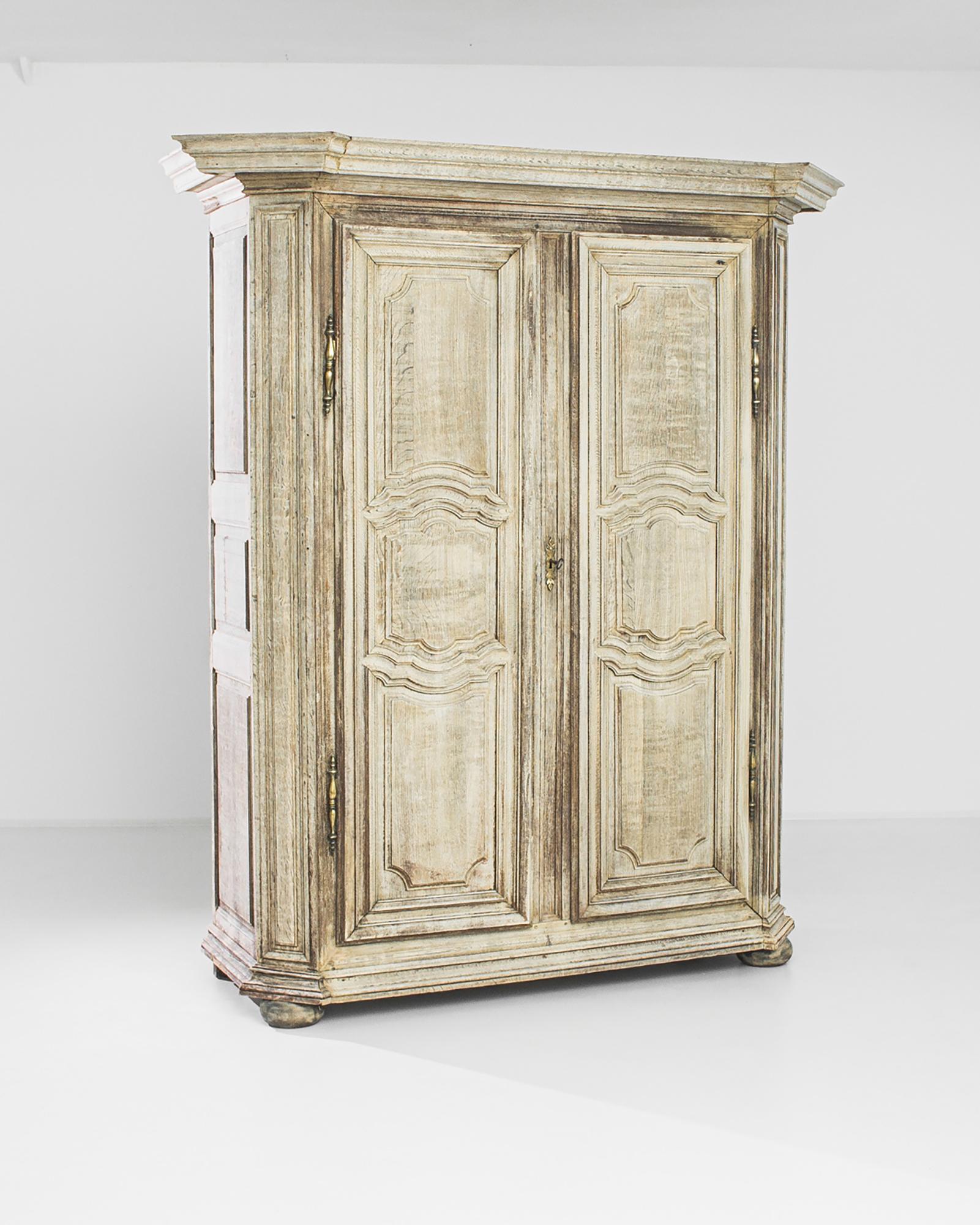 An antique bleached oak cabinet from France, produced circa 1800. A grand wardrobe standing well over seven feet tall, featuring locking double doors with brass hinges and escutcheons. It’s wide base and cornice, in conjunction with the long,