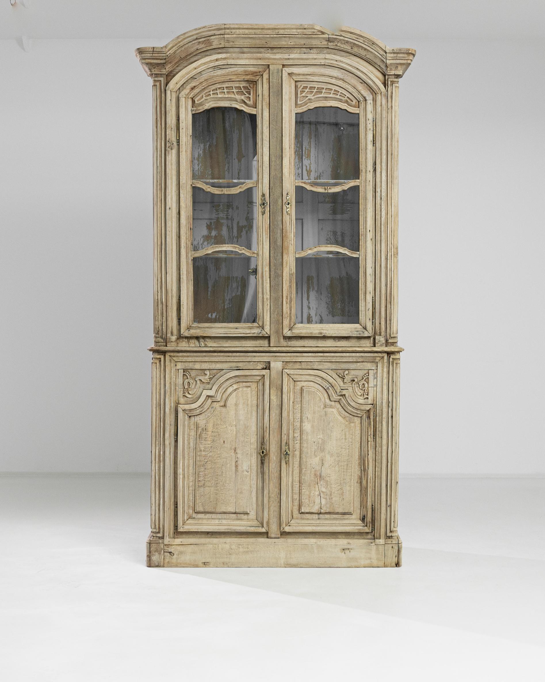 Experience the allure of the 1800s with this French Bleached Oak Vitrine. It is a perfect blend of elegance and functionality. This vitrine has enchanting glass doors at the top that gracefully swing open to reveal two spacious shelves with a