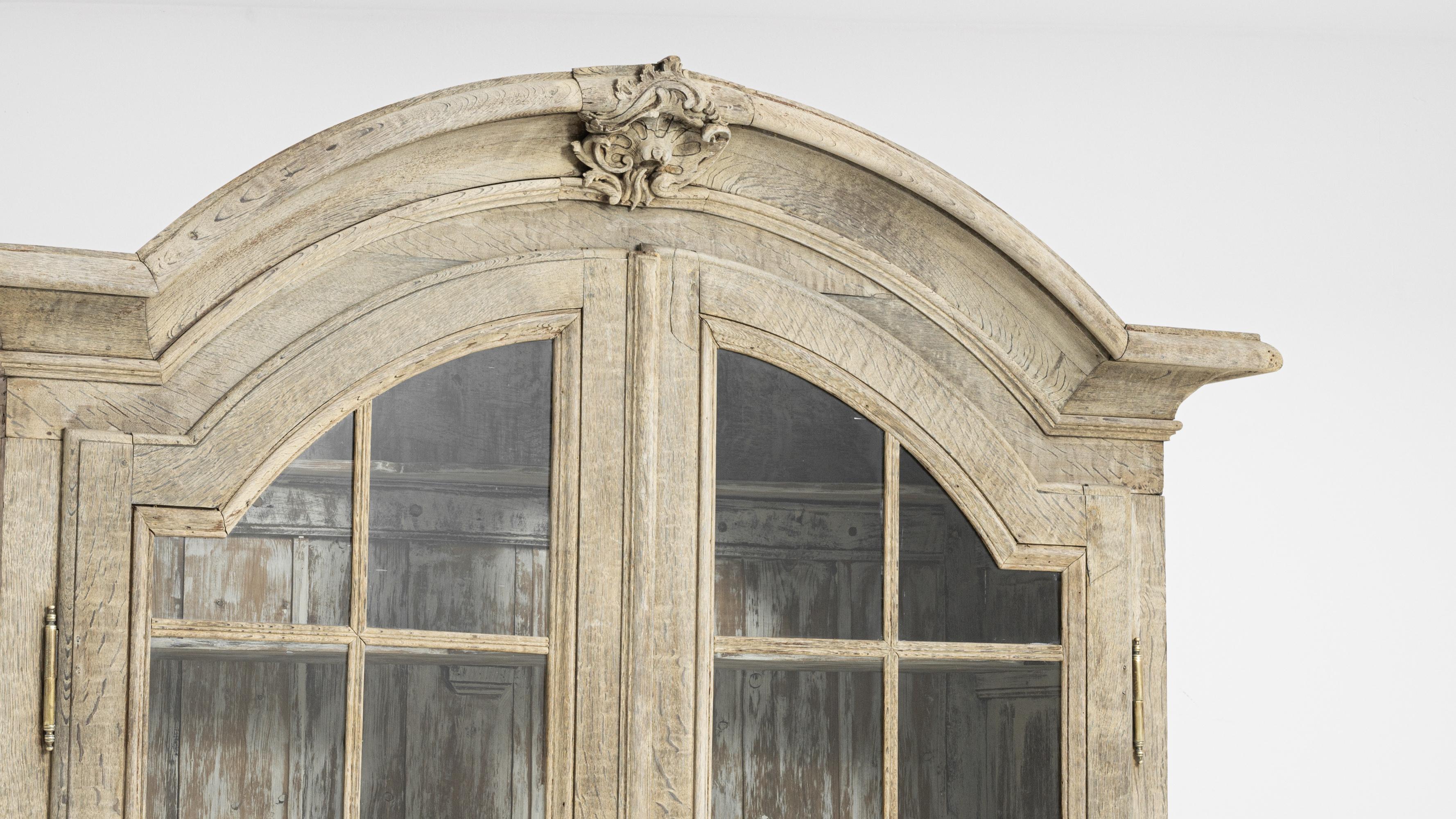 The natural finish of this majestic oak vitrine evokes a view across sand-dunes to the Mediterranean. Built in France, circa 1800, a carved flourish crowns the broad arch of the case — its windswept, sinuous form adds an organic accent to the