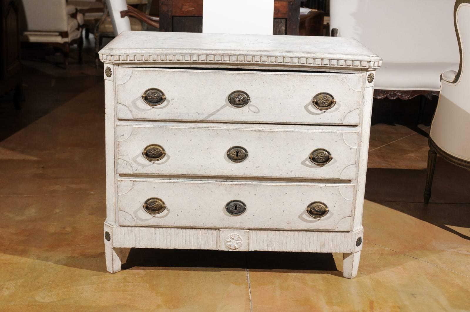 A Swedish period Gustavian painted three-drawer commode from the early 19th century, with dentil molding and rosettes. This Swedish Gustavian chest features a rectangular planked top with canted corners in the front, sitting above a delicate dentil