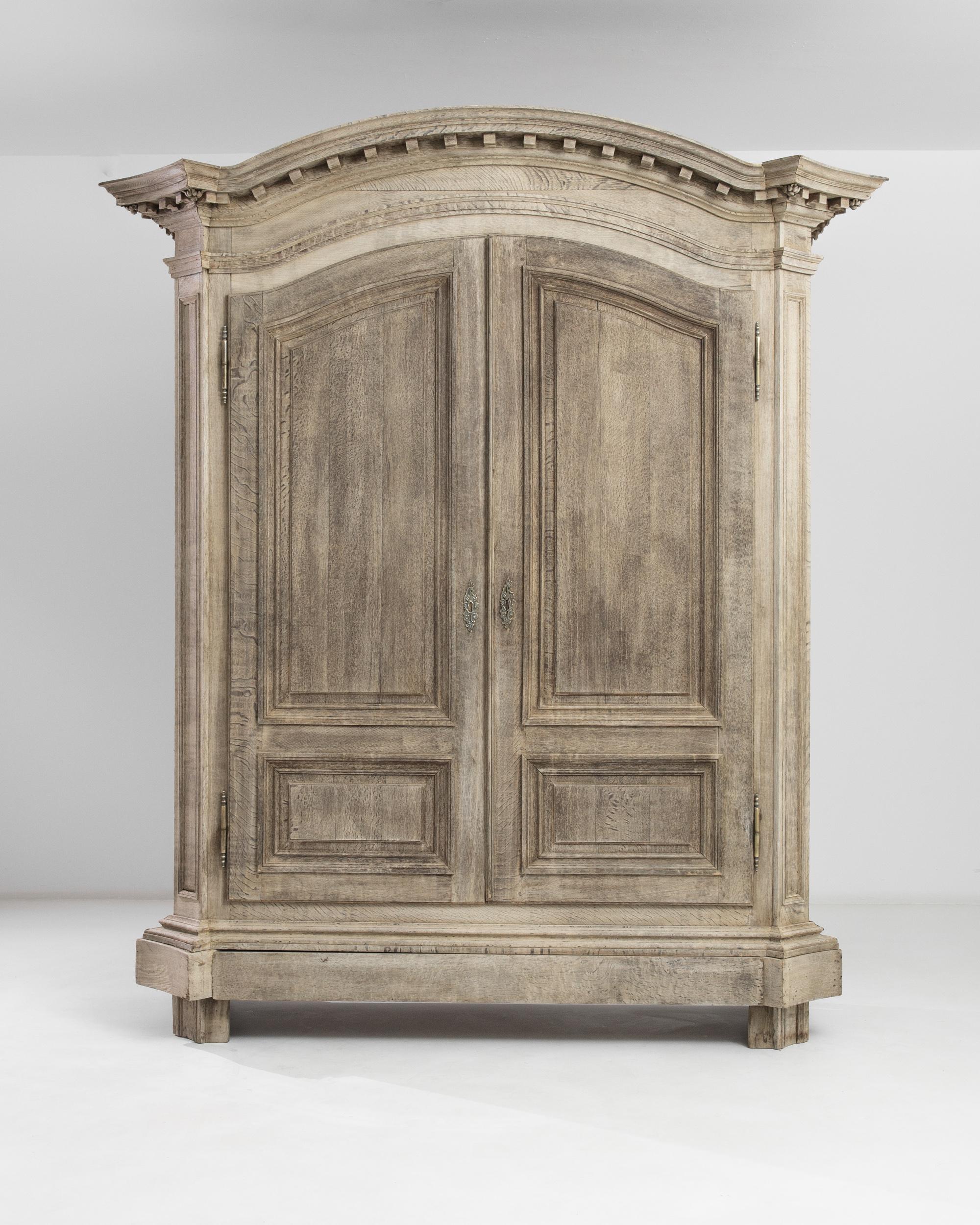 This bleached oak armoire was produced in Belgium, circa 1820. A sturdy case sports three shelves standing on solid bracket feet and cornice base, echoed by a molded top with imposing fortress-like dentils and floral pateras. The tall doors are