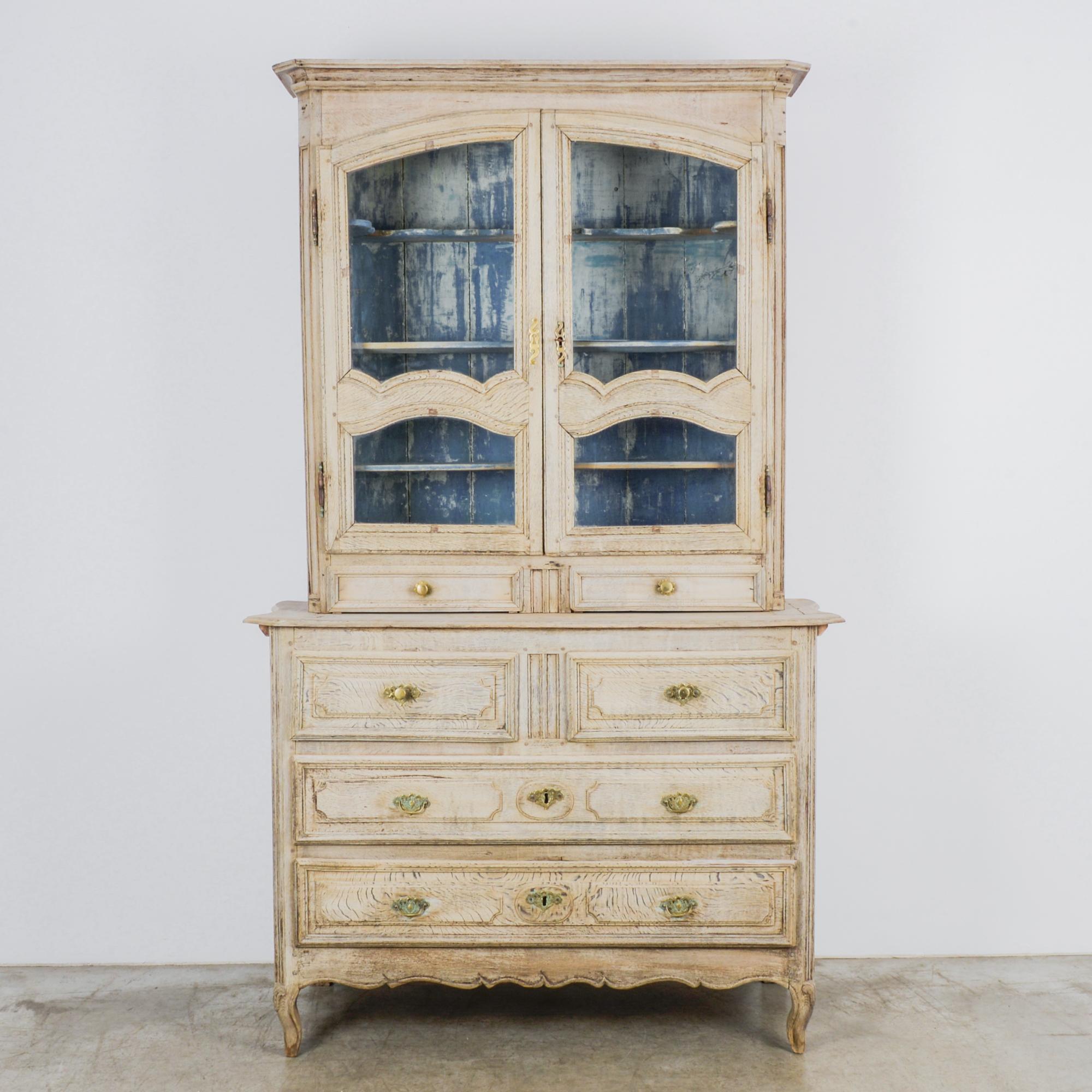 This bleached oak vitrine was made in France, circa 1820. The blue and white interior of the upper section is visible through the panes of the double doors. The lower section houses additional drawers and is raised on cabriole legs. The vitrine