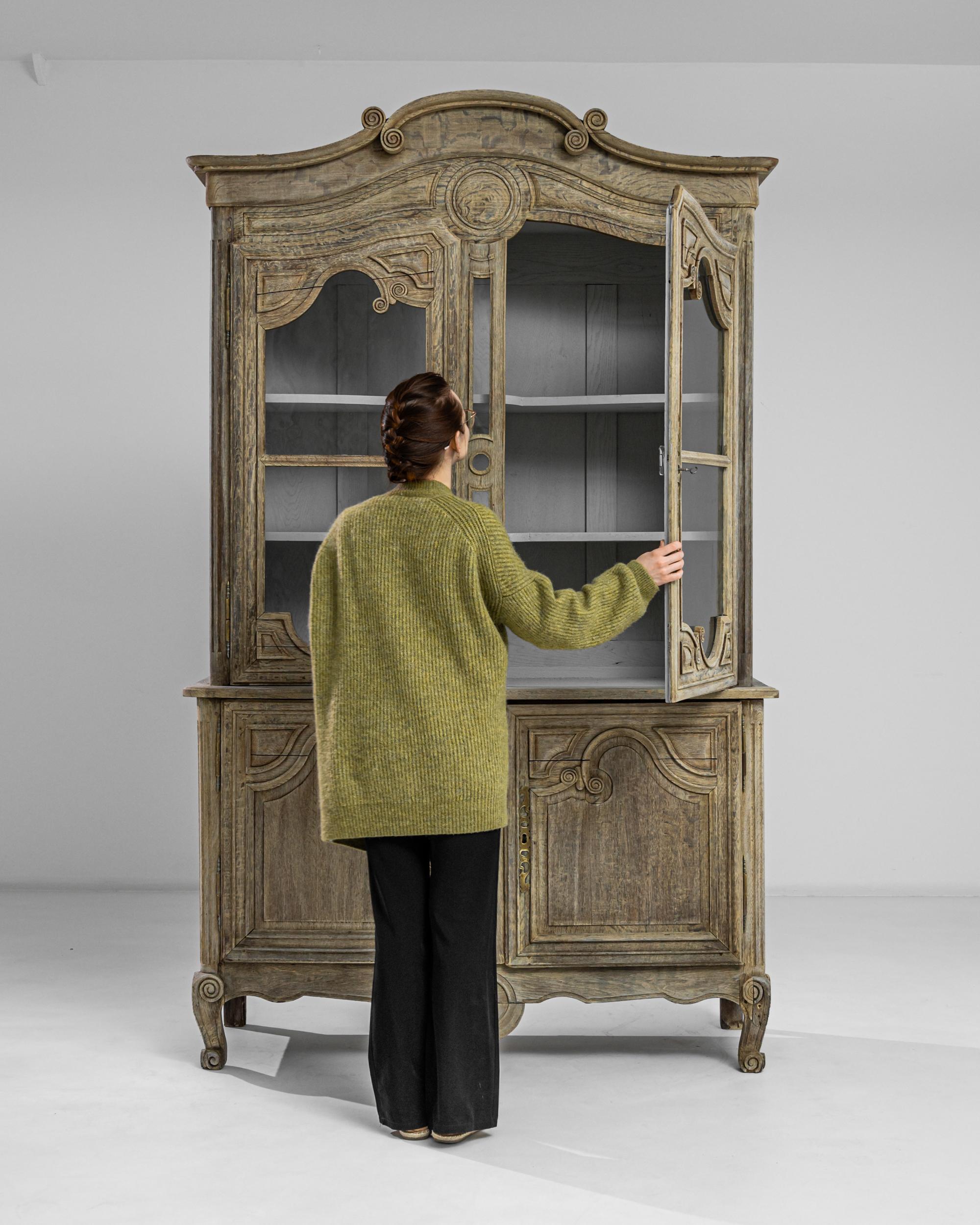 This striking oak vitrine was produced in France, circa 1820. The crafted two-case vitrine stands upon small cabriole feet finished in scrolls, flaunting a scalloped apron and a fully carved front. A multitude of spirals meander around the doors,