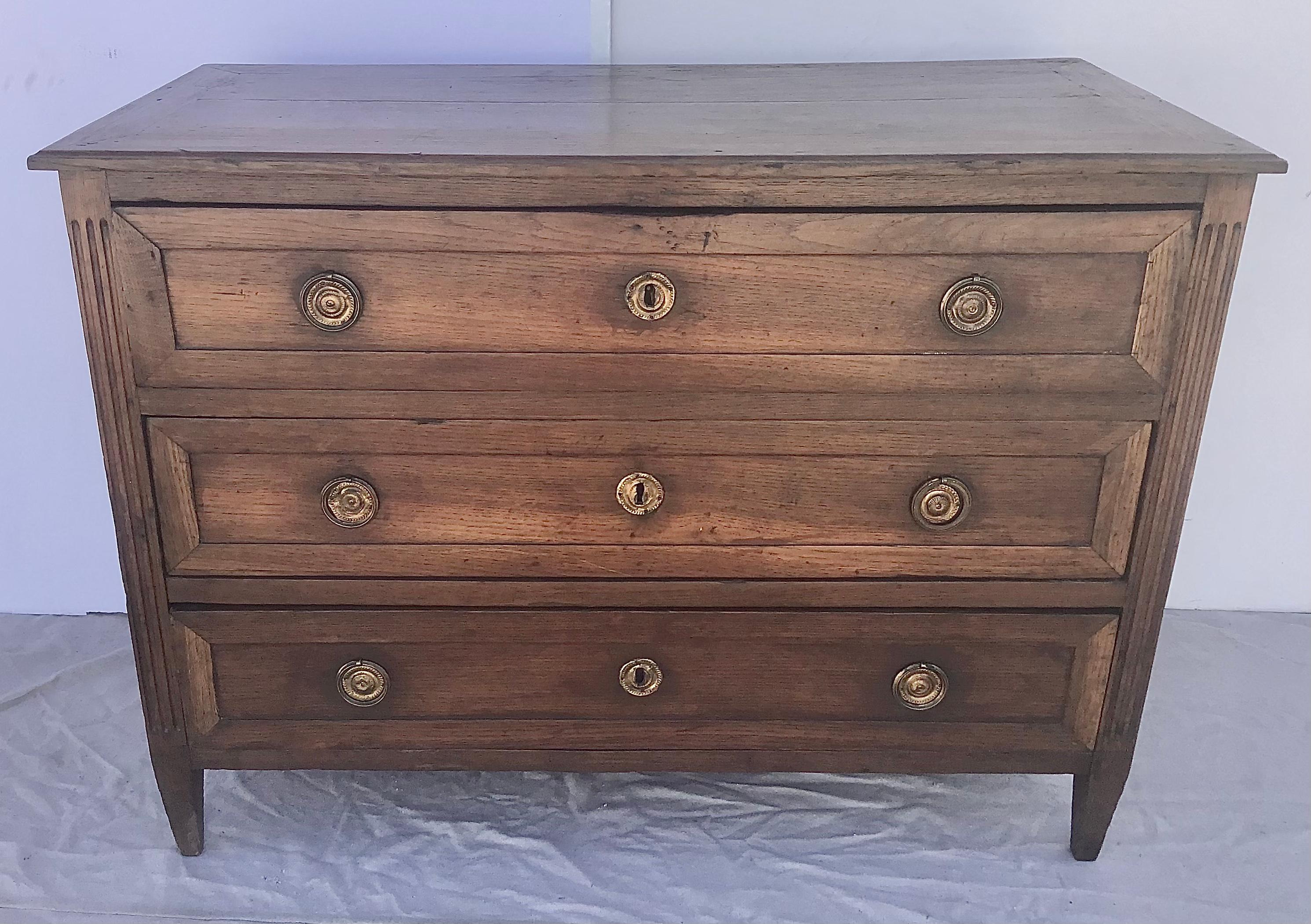 A French neoclassic three-drawer commode with brass trim and tapered feet from the early 19th century. French commode features an exquisite slightly raised rectangular planked top. Three hand-cut dovetailed drawers make up the façade, surrounding