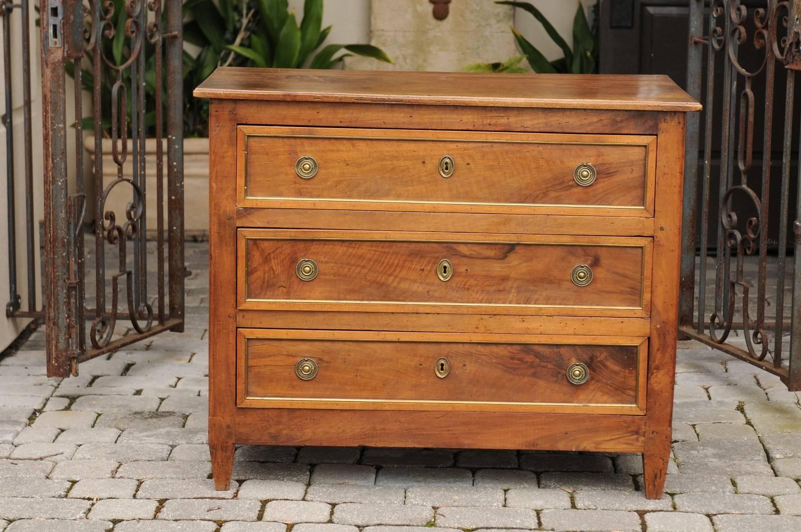 A neoclassical French walnut veneered three-drawer commode with brass trim and tapered feet from the early 19th century. Born in the early years of the tumultuous 19th century, this French commode features an exquisite slightly raised rectangular