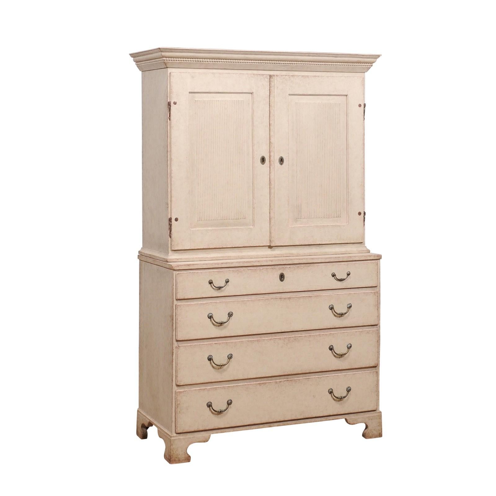 A Swedish two-part tall cabinet from circa 1834 with off white/cream/light grey painted finish, two doors with carved reeded panels, four graduated drawers and carved bracket feet. Step back in time with this exquisite Swedish two-part tall cabinet,