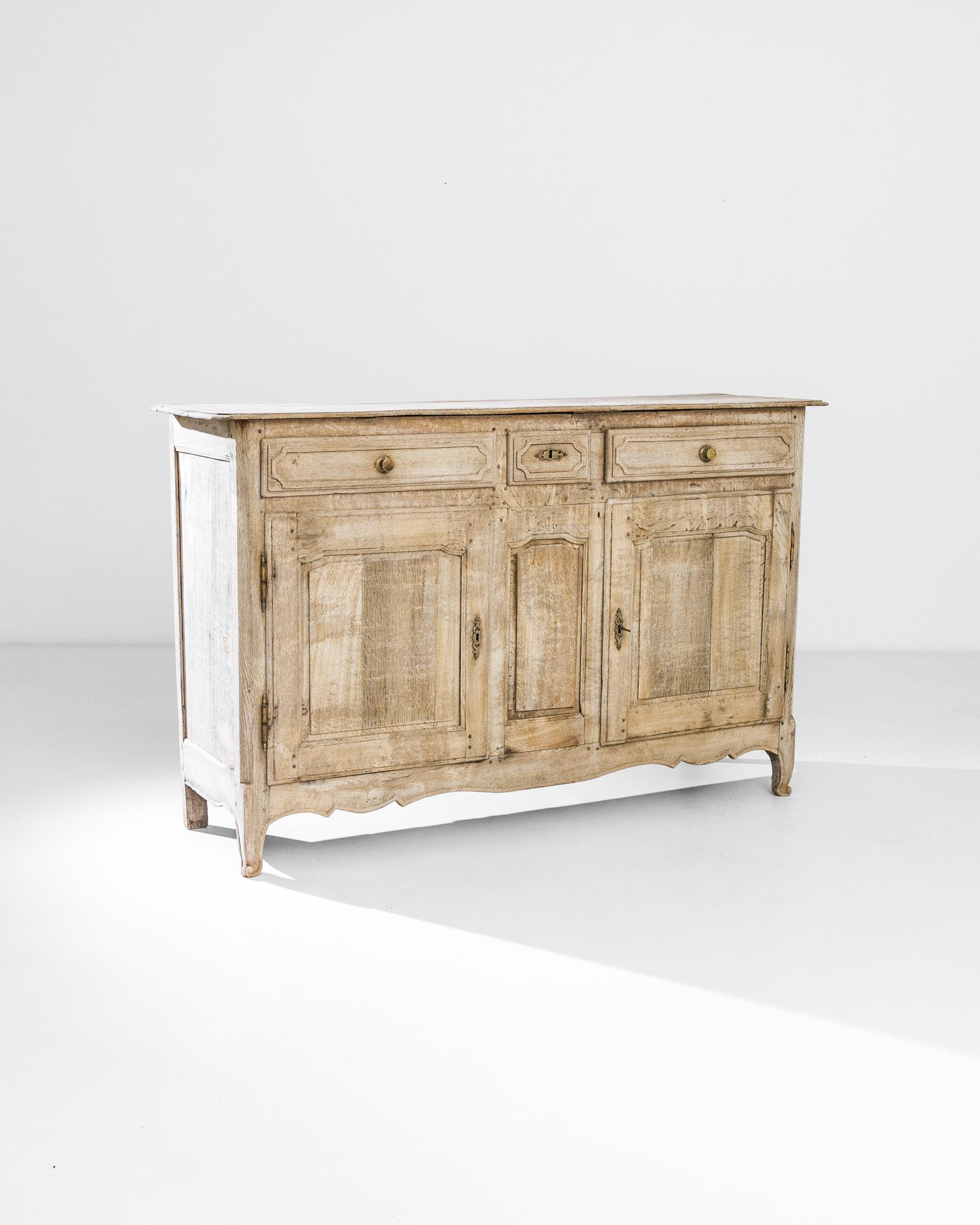 An oak buffet from 1840s France. Out-turned feet, slippered in carved scrolls, give the cabinet a graceful stance. The wood has been restored to natural finish, showcasing the luminous, barleycorn color of the oak, rippled with a subtle grain.