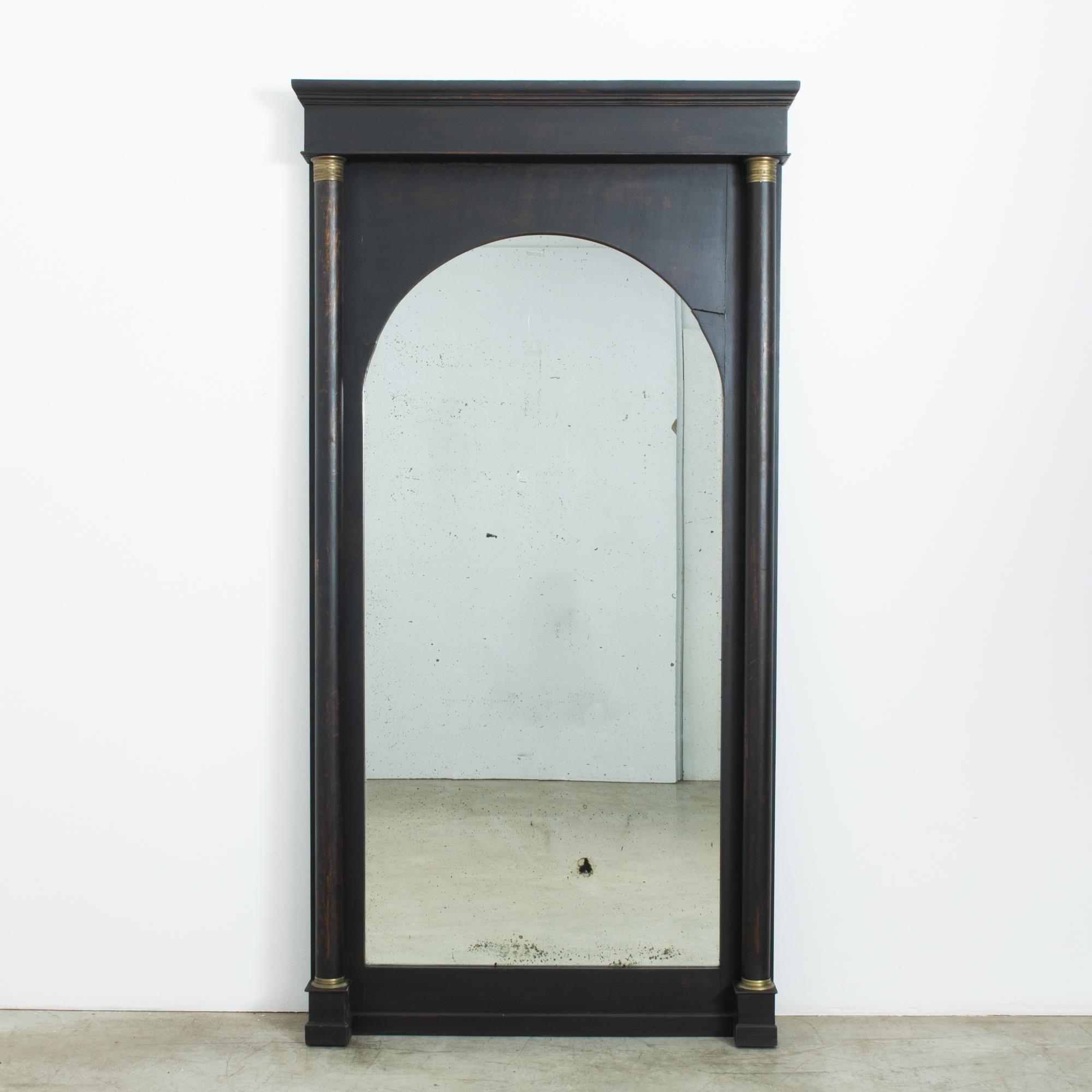 A black, wood framed mirror from France, produced in the 1840s. Standing at an impressive six feet six inches, this tall mirror stretches into a smooth arch. An Empire influneced style, flanked by two gold-capped pillars. Now delicately spotted with