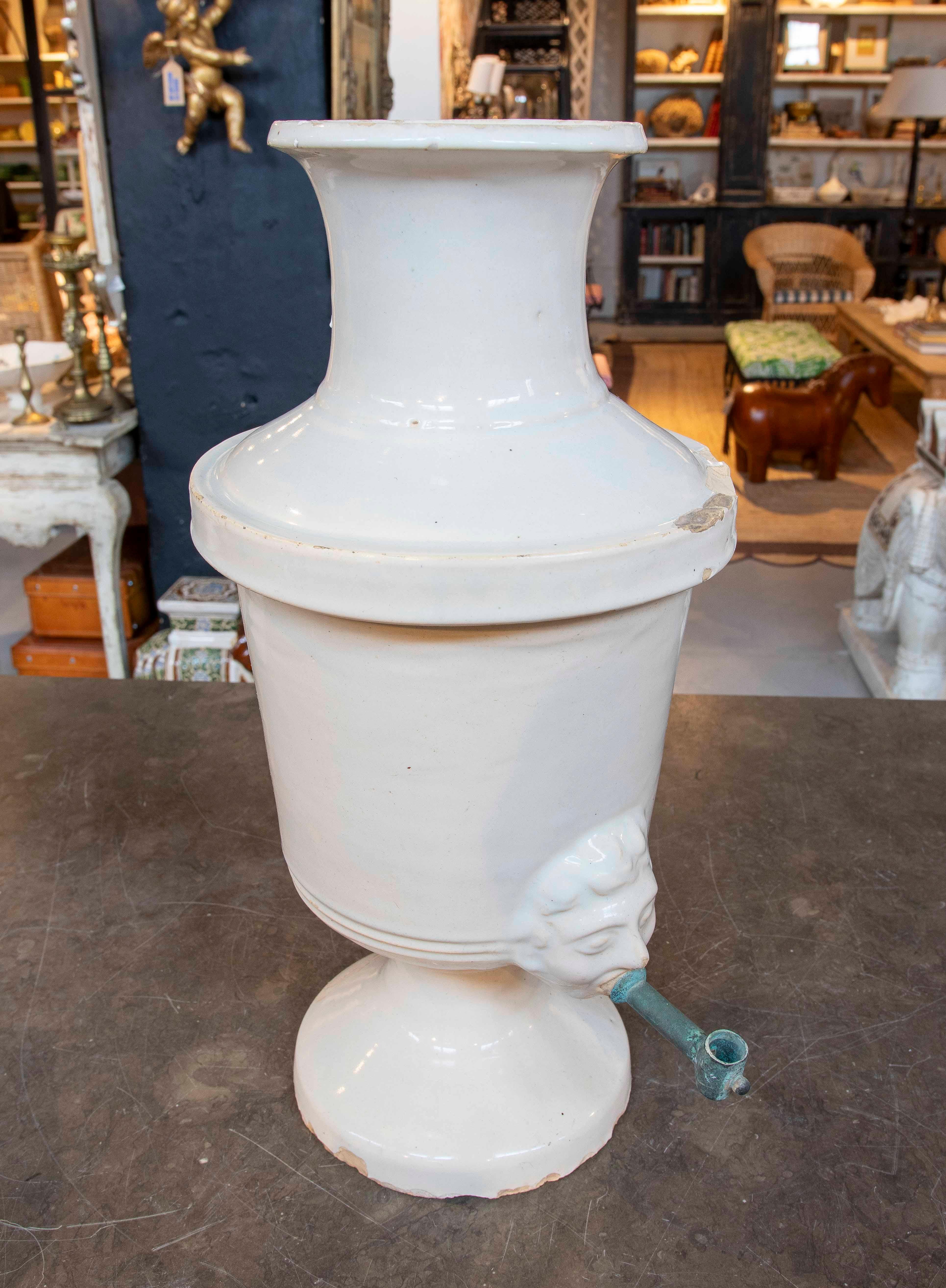 Elegante e unico dispenser in ceramica smaltata bianca, con dettagli ornamentali che includono un volto scolpito in rilievo da cui emerge il rubinetto in metallo. Il pezzo ha un design classico con base a piedistallo e collo svasato. La patina e
