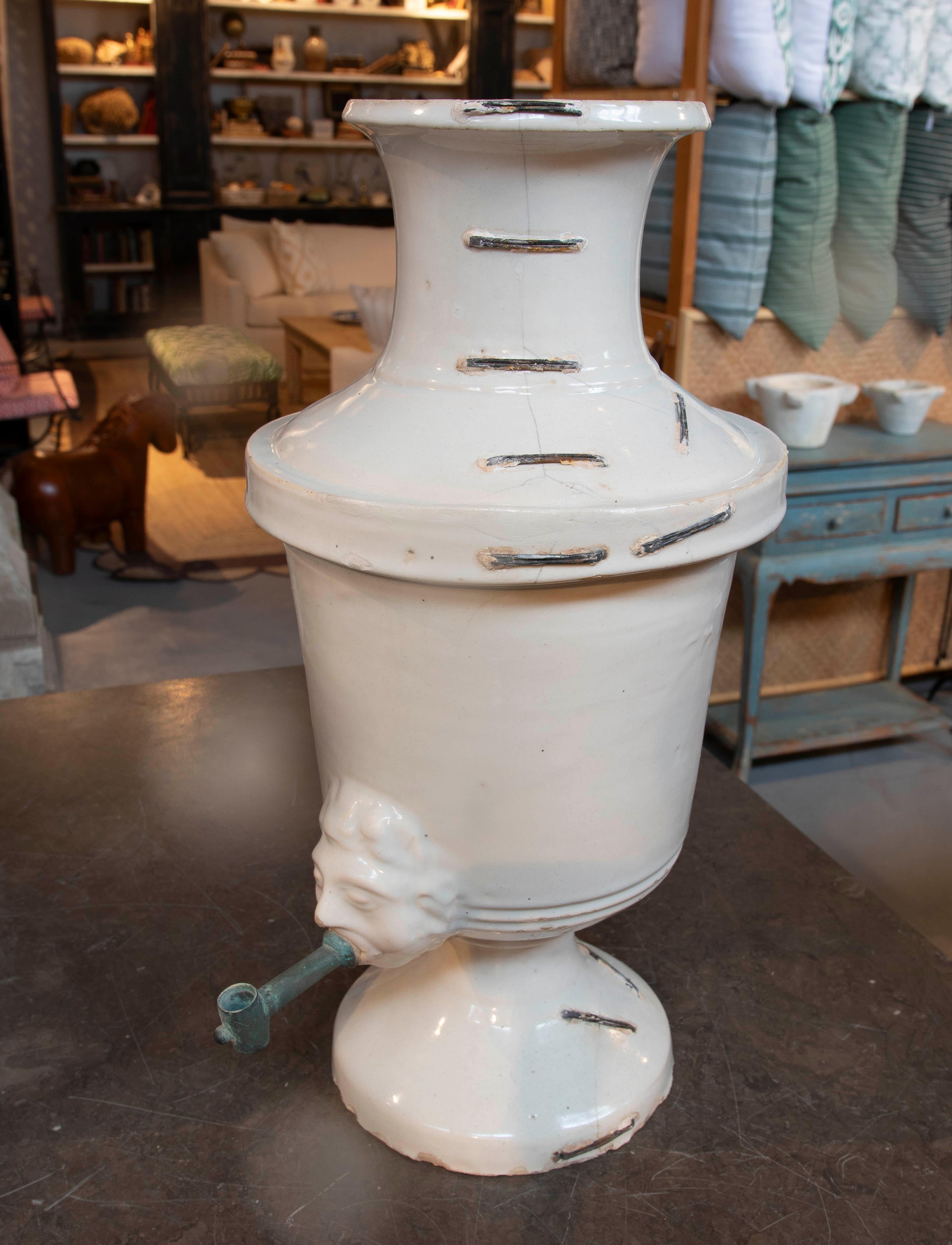 Distributeur élégant et unique en céramique émaillée blanche, avec des détails ornementaux qui incluent un visage sculpté en relief d'où émerge le robinet en métal. La pièce présente un design classique avec une base piédestale et un col évasé. Sa