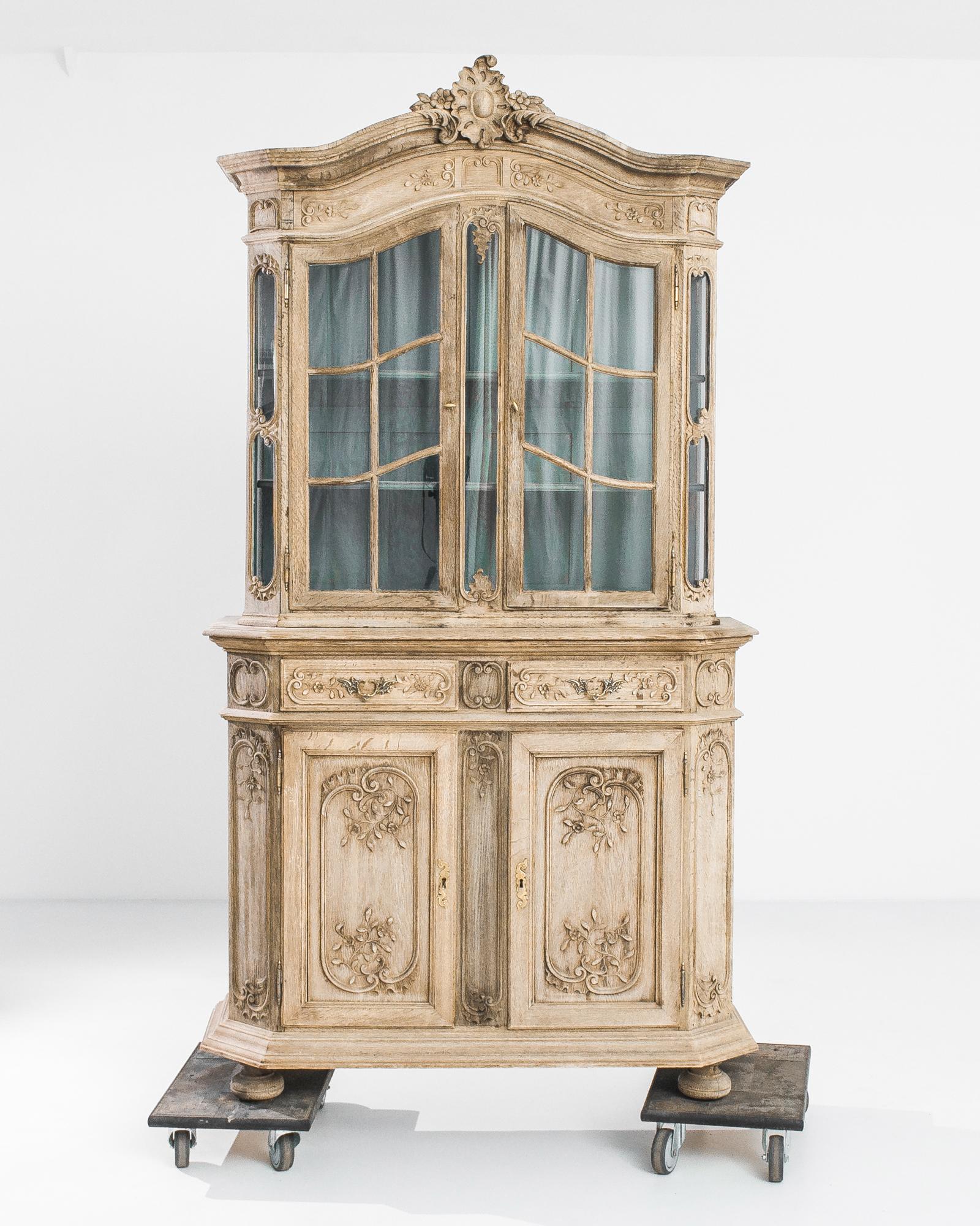 A bleached oak vitrine from Belgium, produced circa 1850. A stately display cabinet that stands like a sculpture in the round, featuring a three shelf upper cabinet, two central siding drawers, and a lower cabinet of two shelves. Filled with