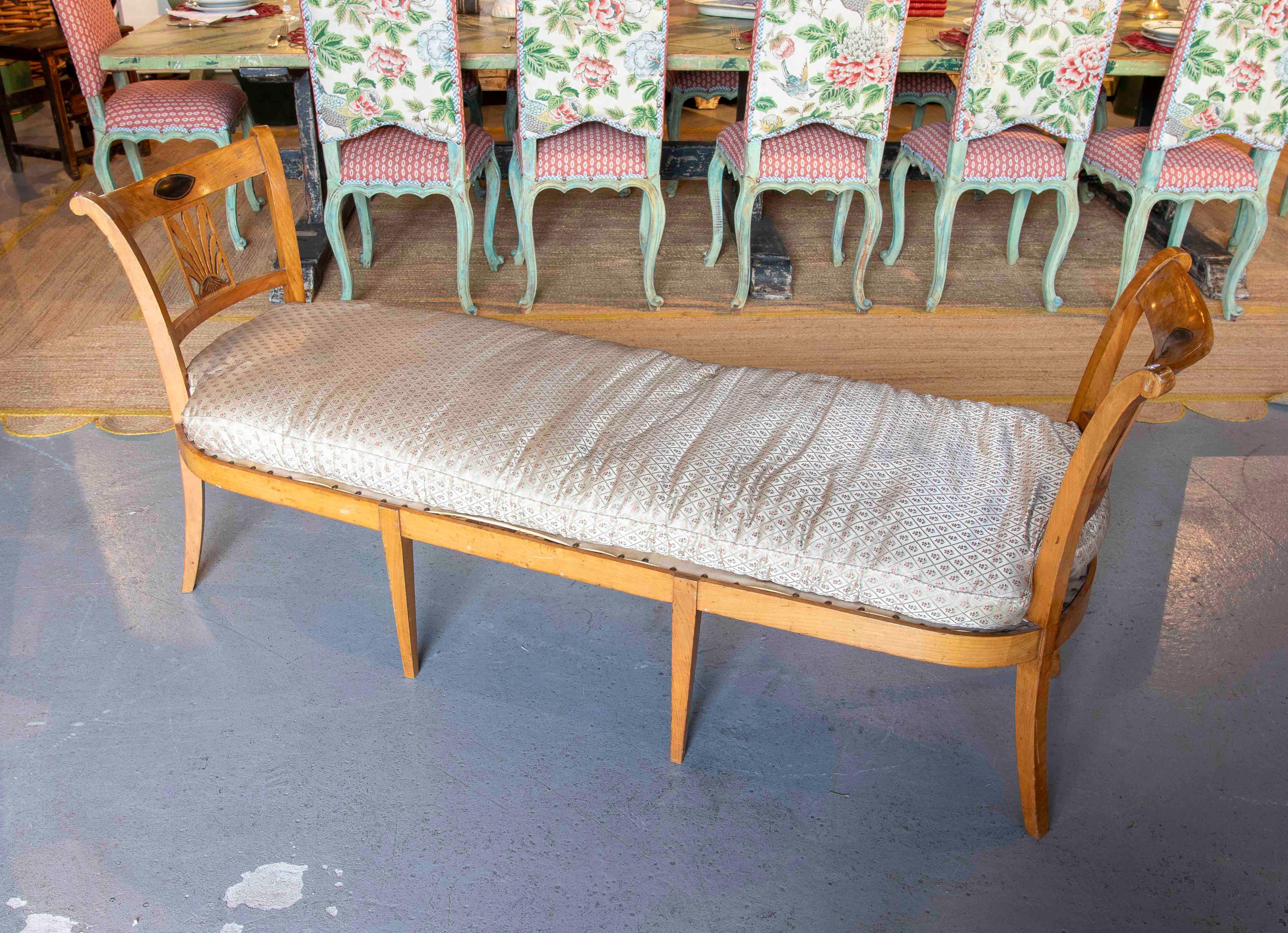 Elongated bench made from Bierdermeier wood with a design that includes a backrest at both ends. The structure features simple lines that provide functionality and support. It includes an upholstered cushion in geometric patterned fabric that adds