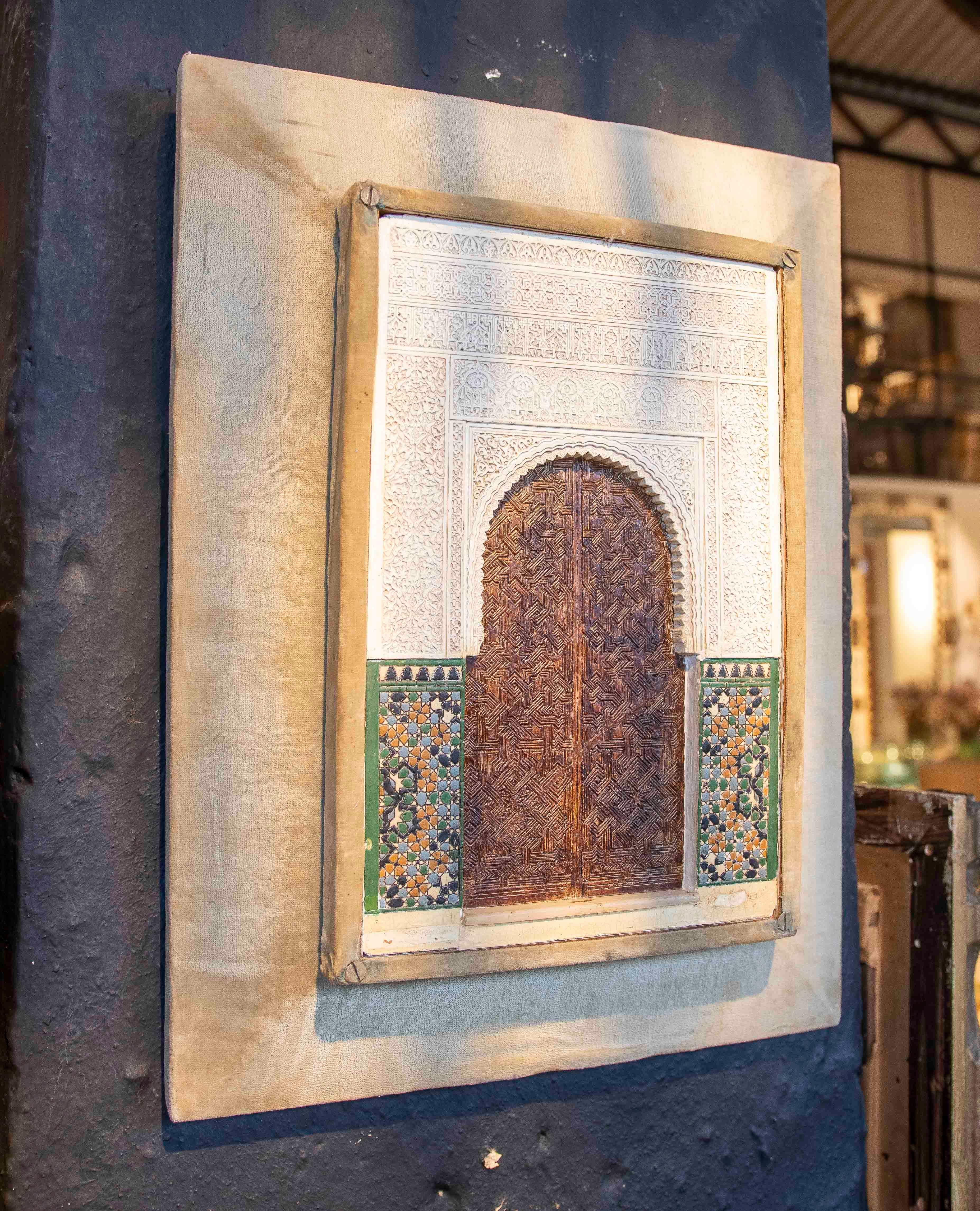 Magnifique maquette murale reproduisant une porte en arc en fer à cheval, minutieusement sculptée dans le bois selon un motif géométrique typique de l'art mudéjar. La porte est en stuc avec de délicates inscriptions et des motifs en treillis, tandis