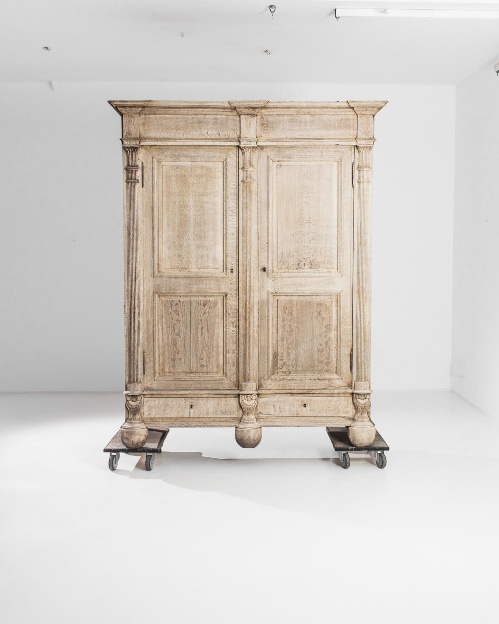 An oak cabinet from 1850s France with a stately neo-classical aesthetic. Paneled doors are framed by fluted Egyptian columns, crowned by a capital of fanning palm leaves. At the base, carved victory urns, wreathed with laurel, sit atop stocky