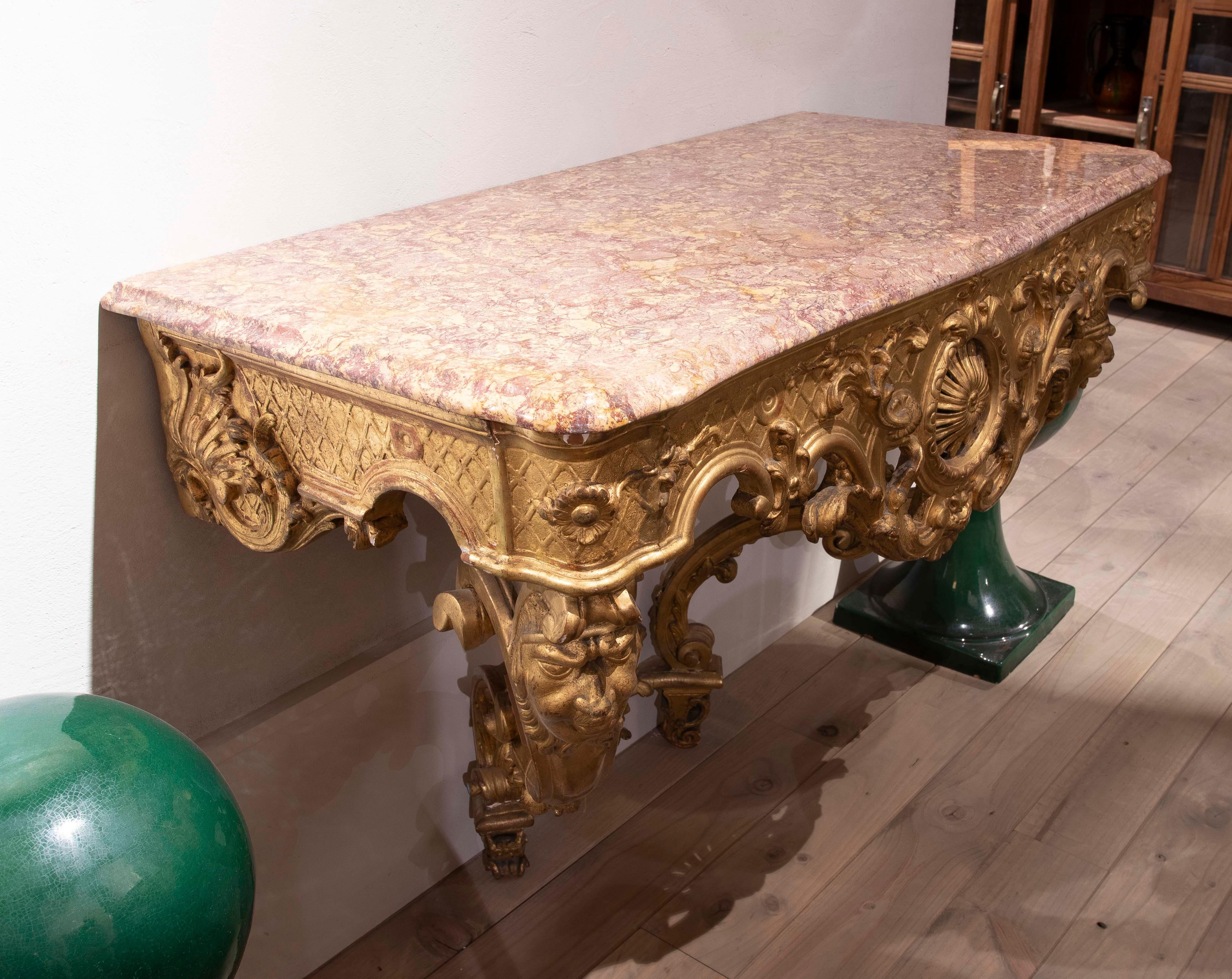 An exquisite Baroque-style console table, richly carved in wood and finished in gold leaf, with an imposing veined pink marble top. The structure displays an exuberant and dynamic design, with ornate details such as scrolls, acanthus leaves, shells,