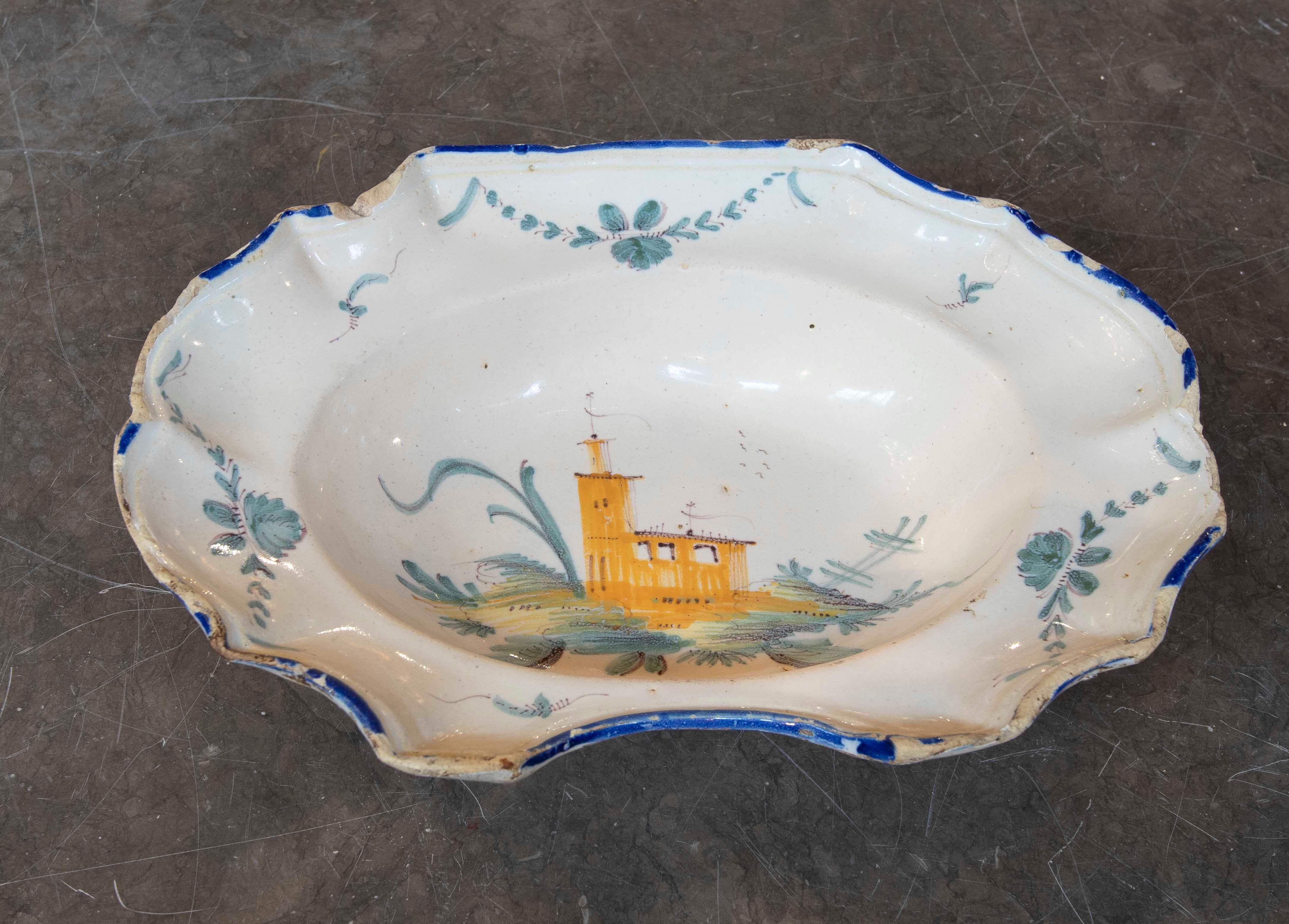 Plato oval de cerámica esmaltada, decorado a mano con una escena paisajística central en la que aparece una torre o castillo amarillo rodeado de elementos naturales estilizados. El borde está adornado con motivos florales en tonos azules y verdes,